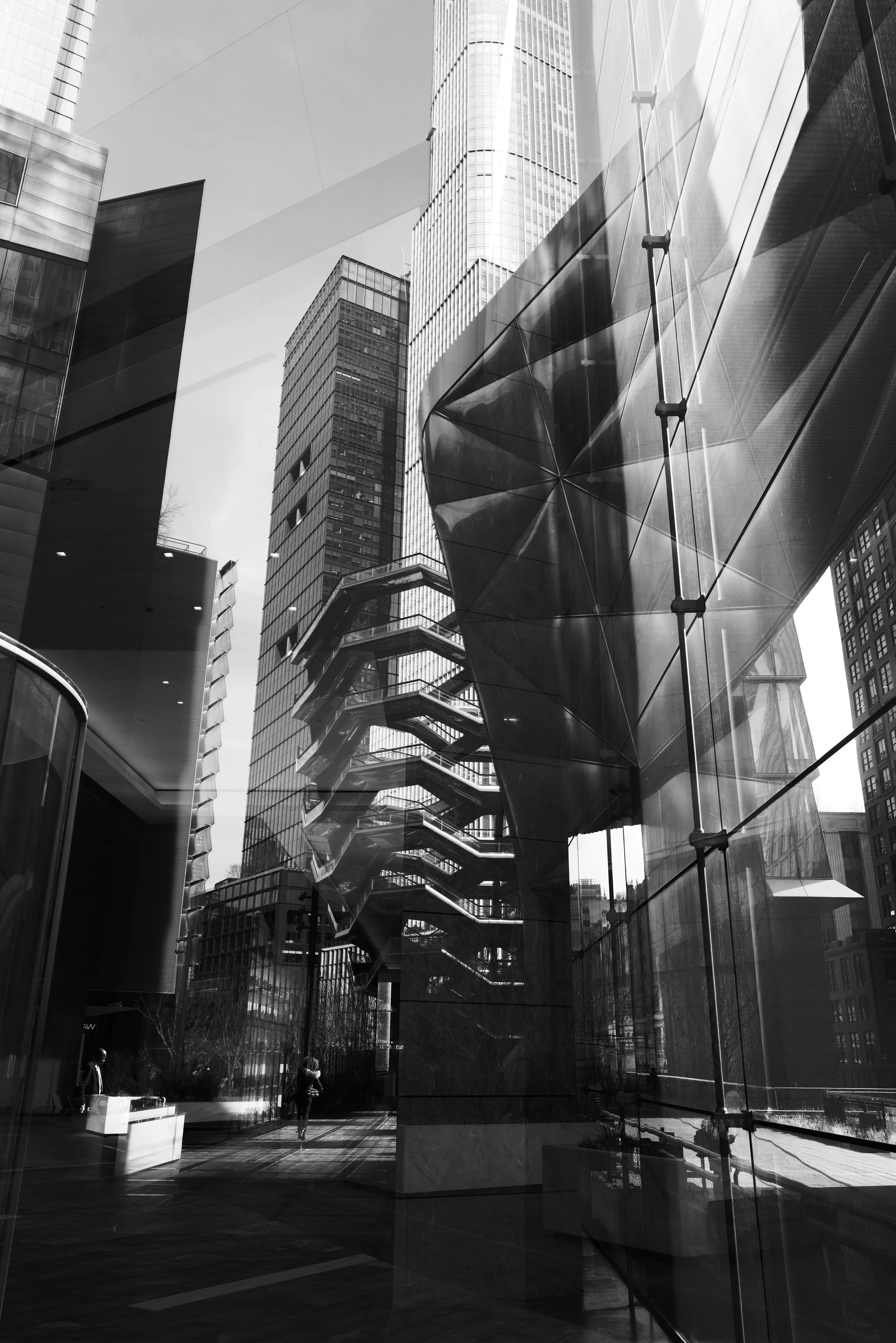 Black and white photo of a modern cityscape with tall glass and steel buildings, including a distinctive angular building with external fire escape staircases.
