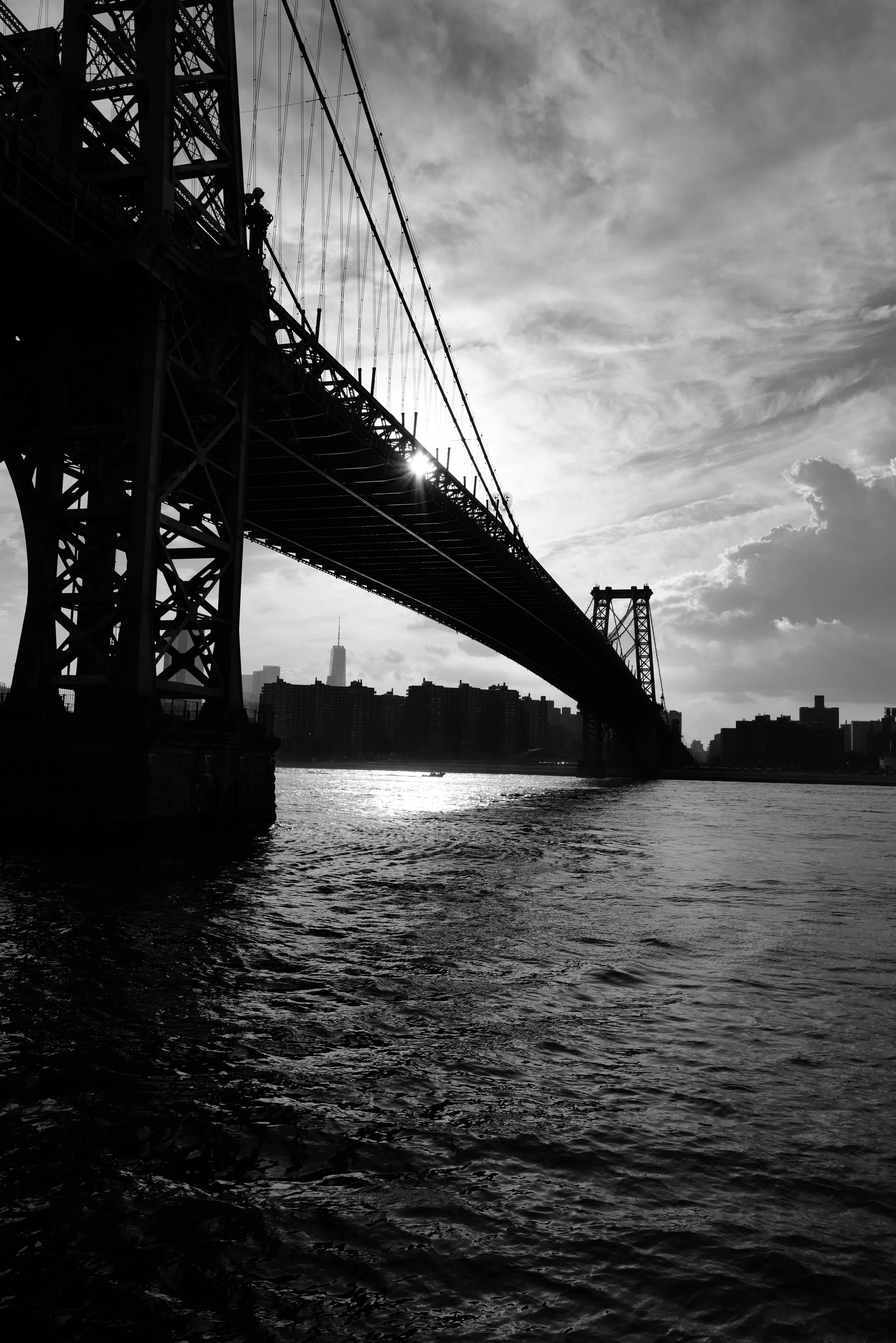 Black and white photo of a bridge over a body of water with a city skyline in the background, including a famous skyscraper, during sunset or sunrise.