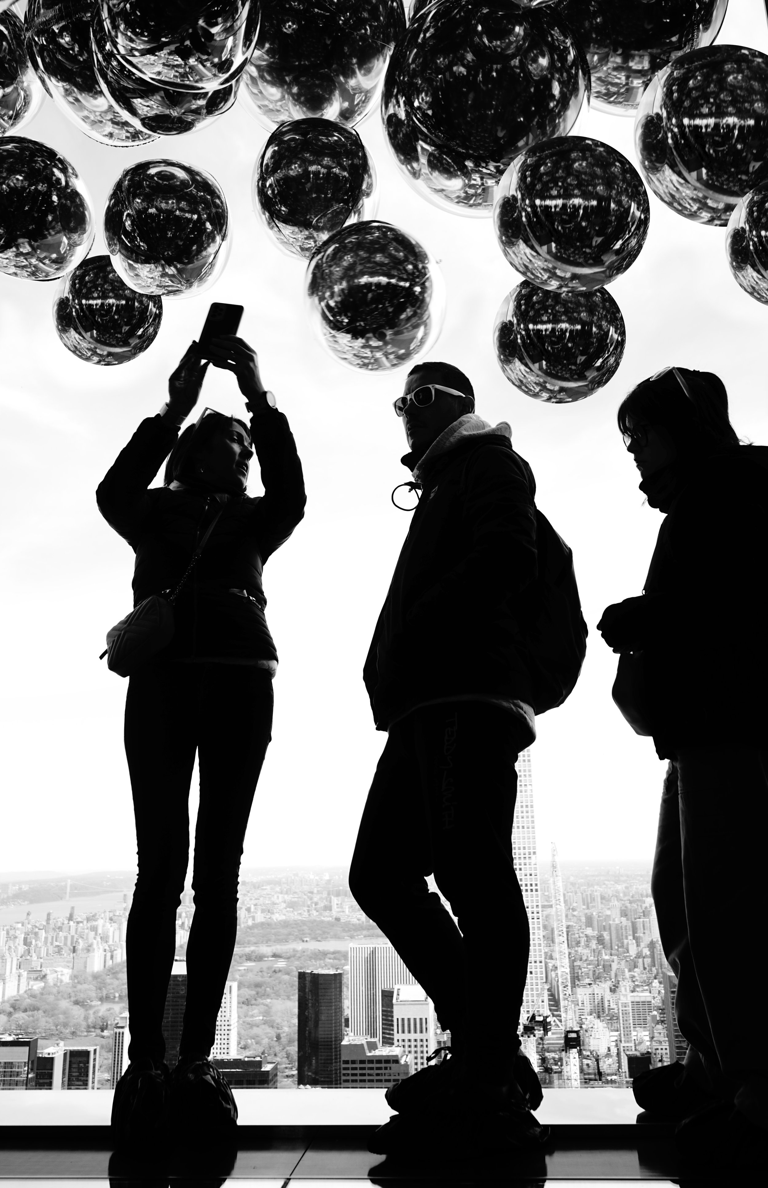 Silhouettes of three people standing on a high rooftop overlooking a city skyline, with balloons above and cloudy sky.