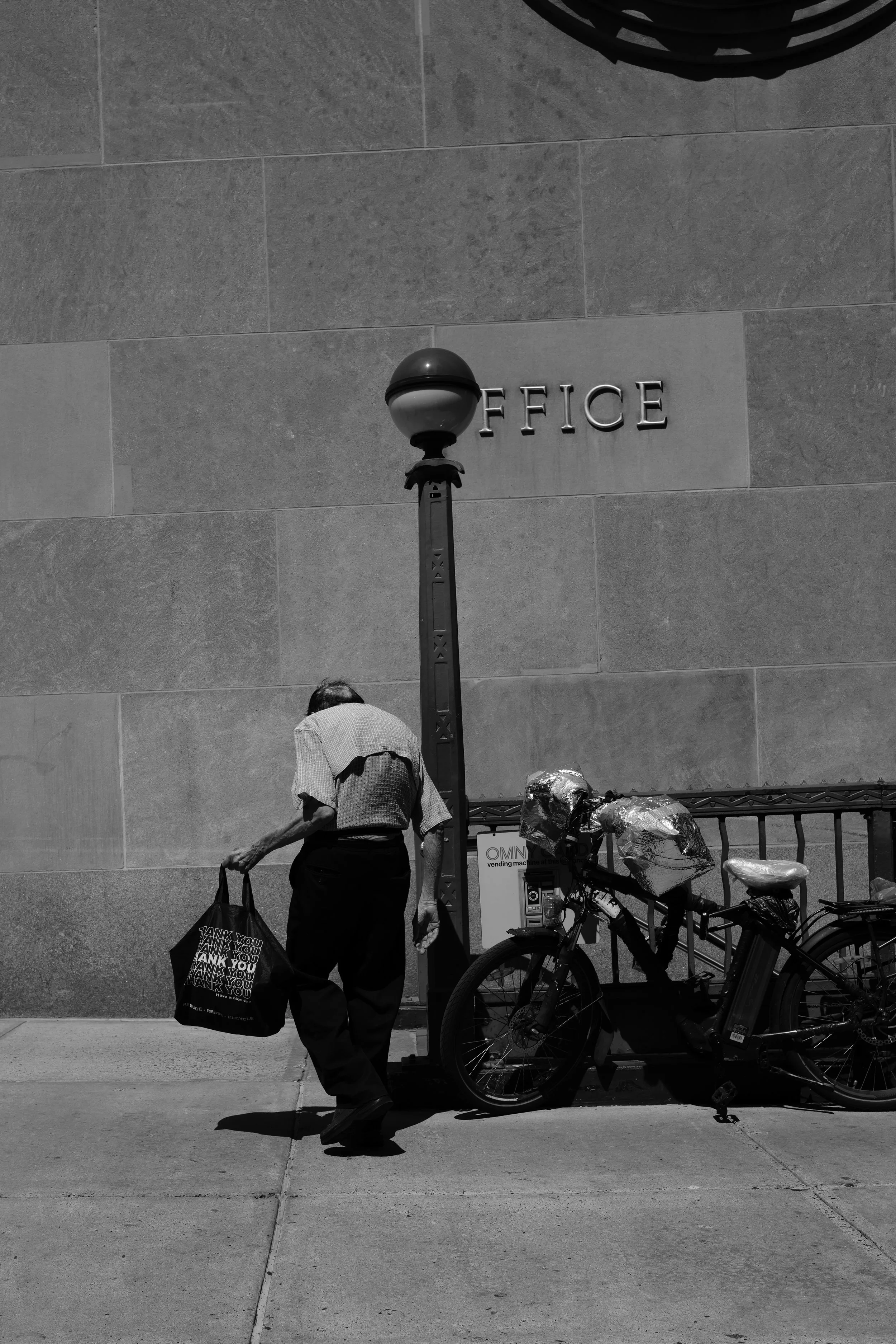 An elderly person walking past a parked bicycle with plastic bags on it, on a sidewalk in front of a building with a sign that says 'OFFICE' and a street lamp.
