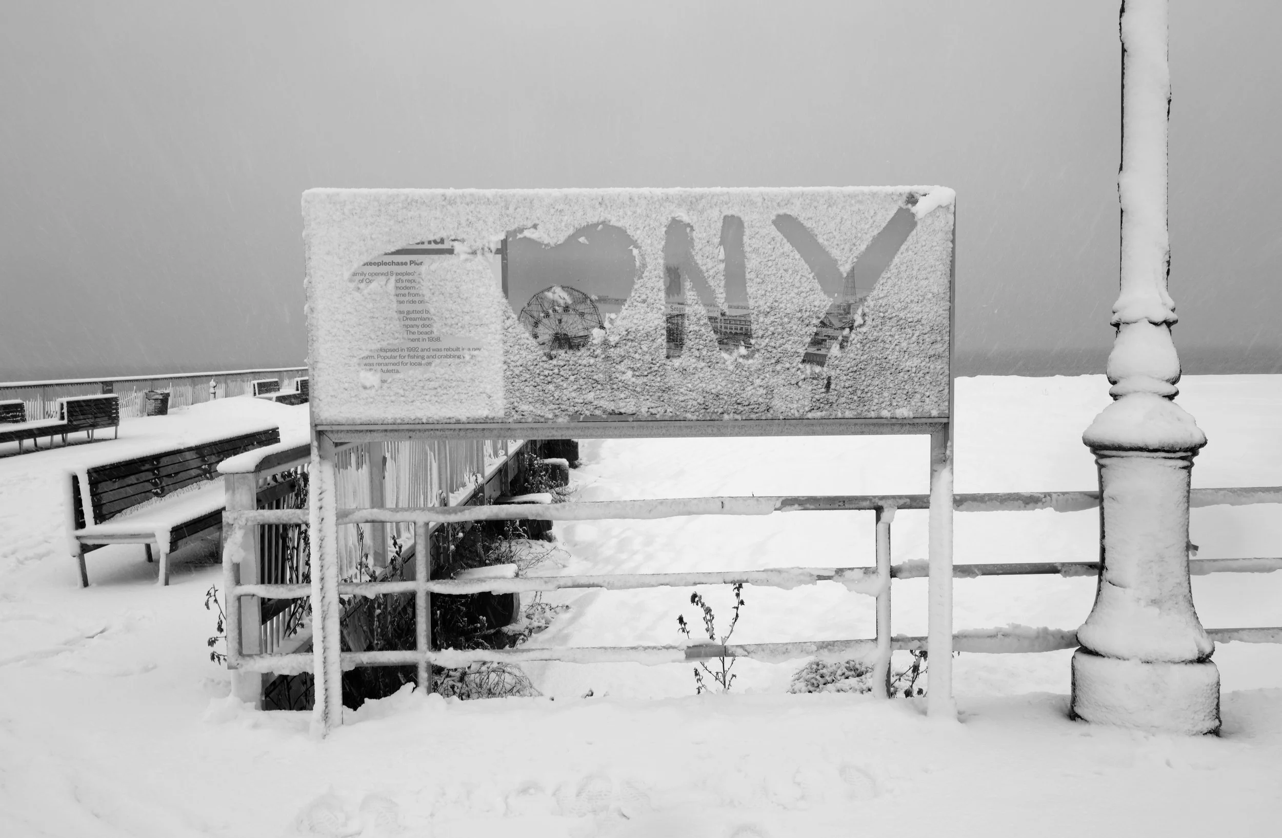 Snow-covered sign with the words 'TOMY' partially obscured, on a snow-covered pier with benches and a snowy landscape in the background.