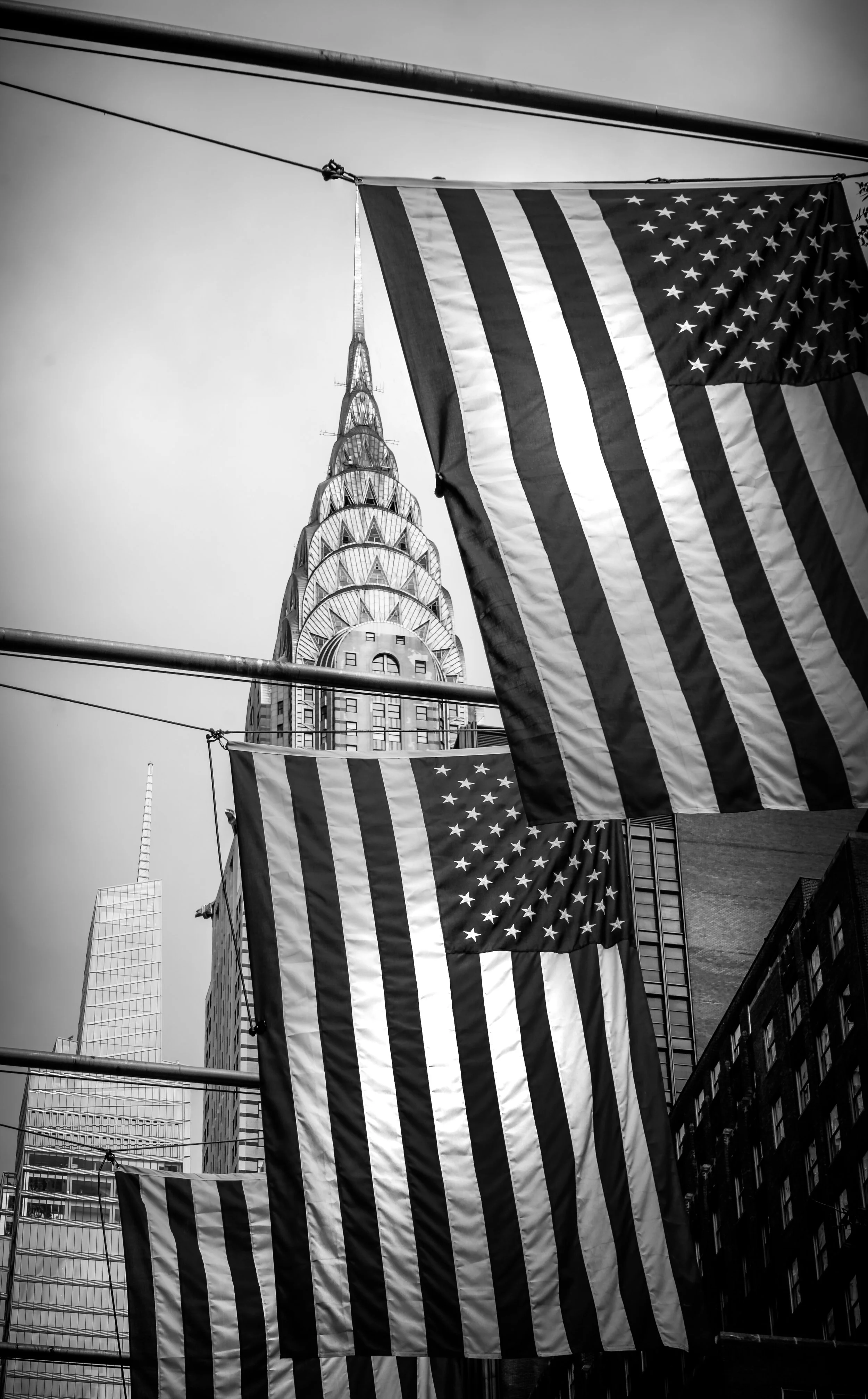 Black and white photo of American flags hanging between city buildings, with the Chrysler Building in the background.