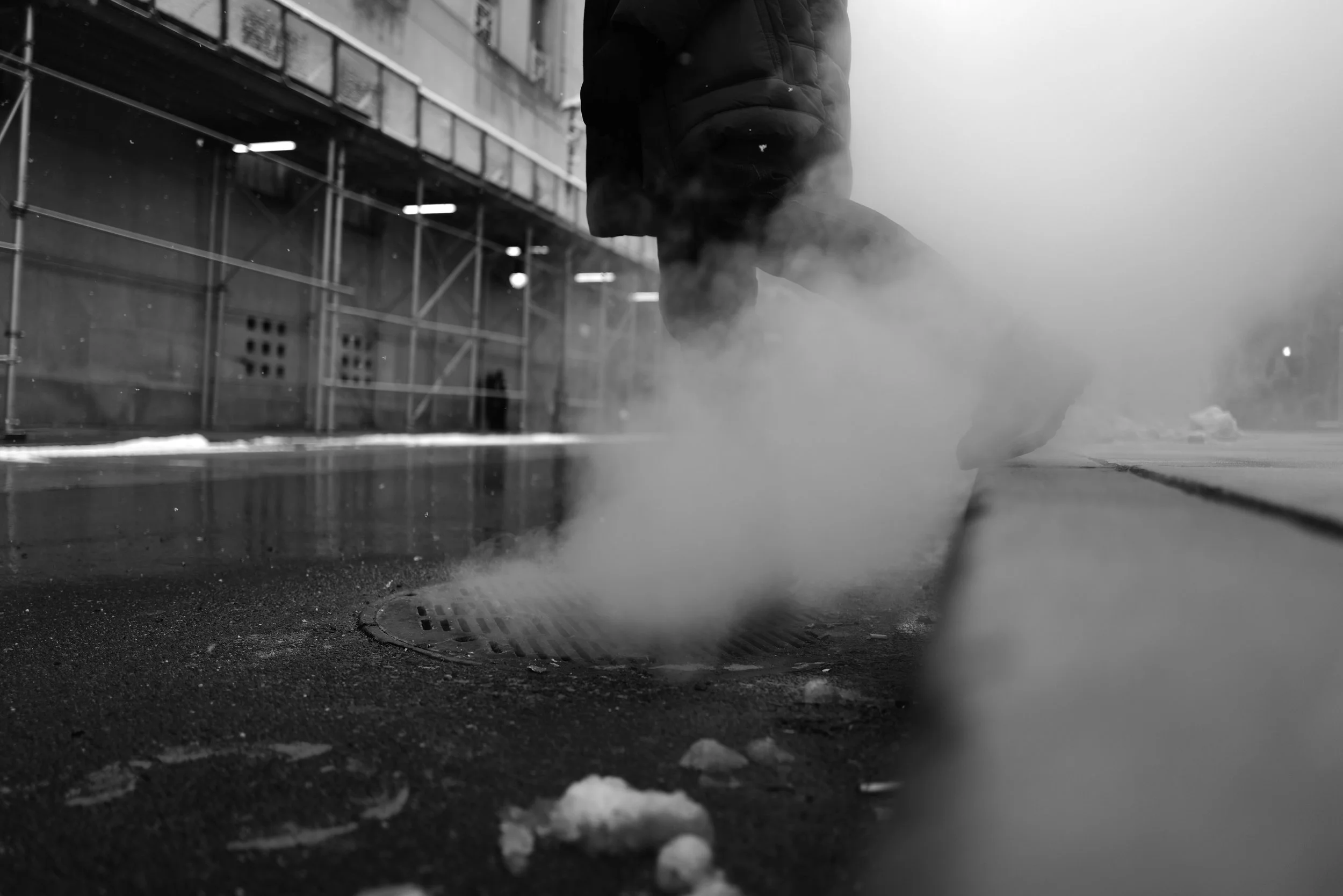 A person walking past a steaming manhole cover on a city street in black and white.