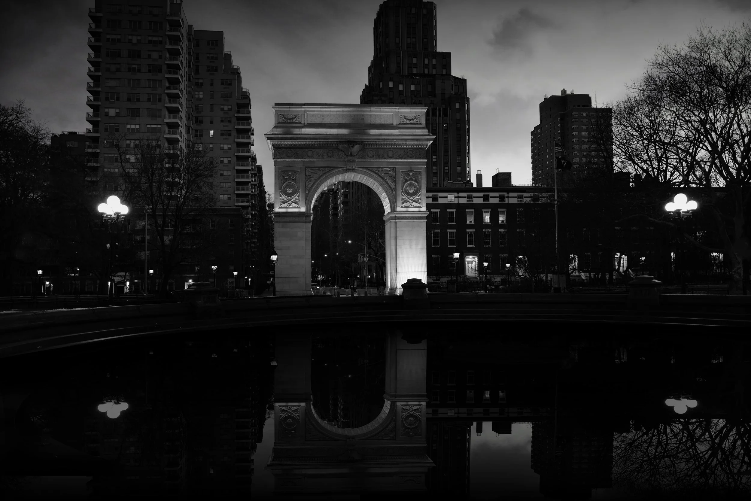 Black and white photo of Washington Square Arch illuminated at night, with tall buildings and leafless trees in the background, and their reflection in a water feature in the foreground.