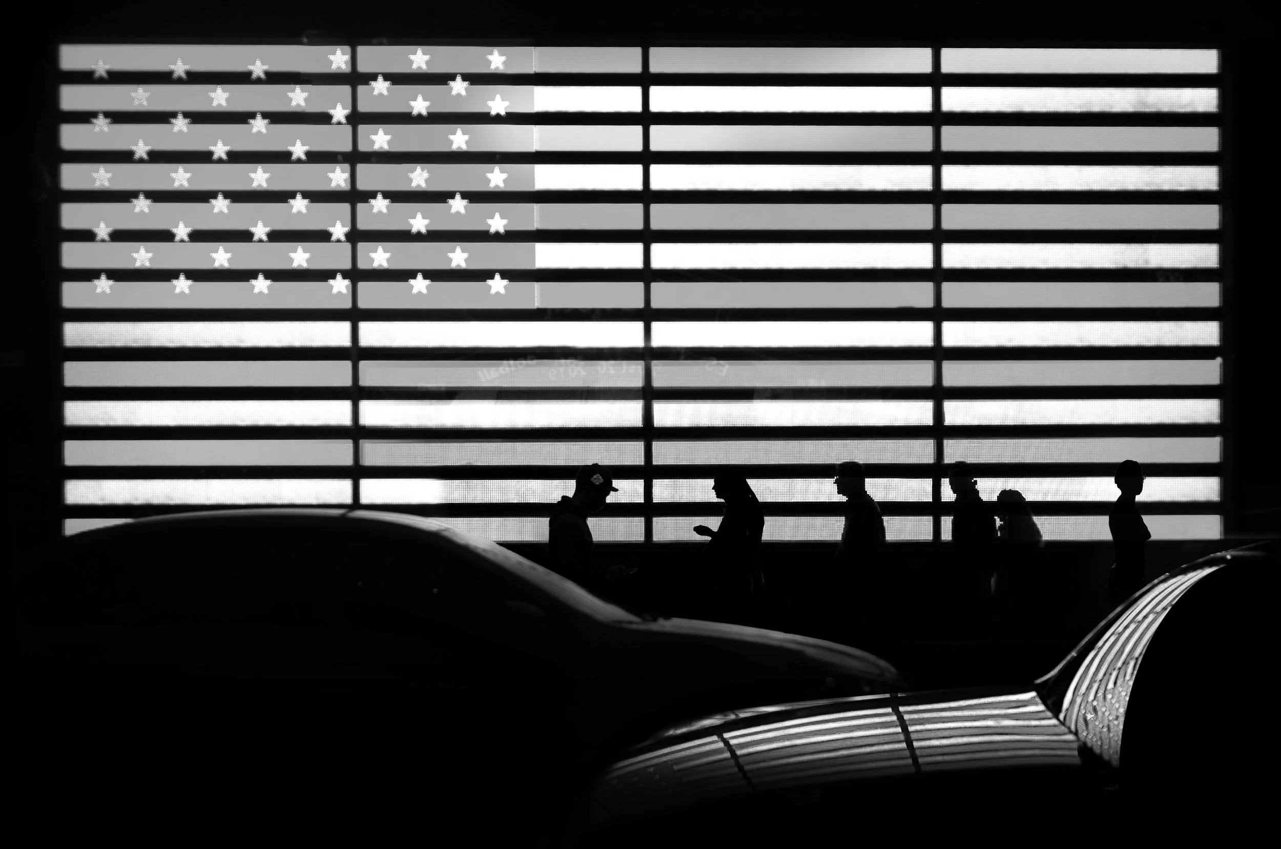 Silhouettes of five people walking indoors in front of a large American flag display, with cars in the foreground.