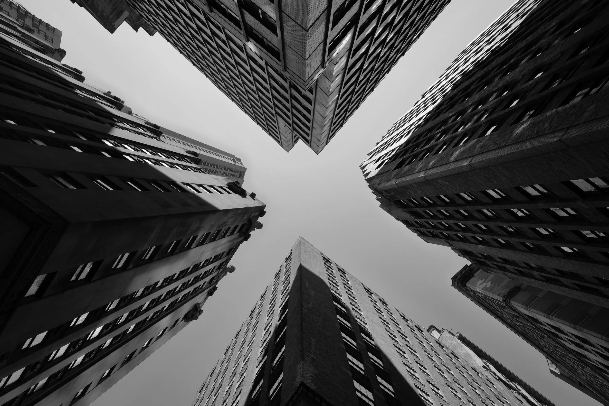 Black and white photo of four tall skyscraper buildings taken from ground looking up, converging towards the sky.