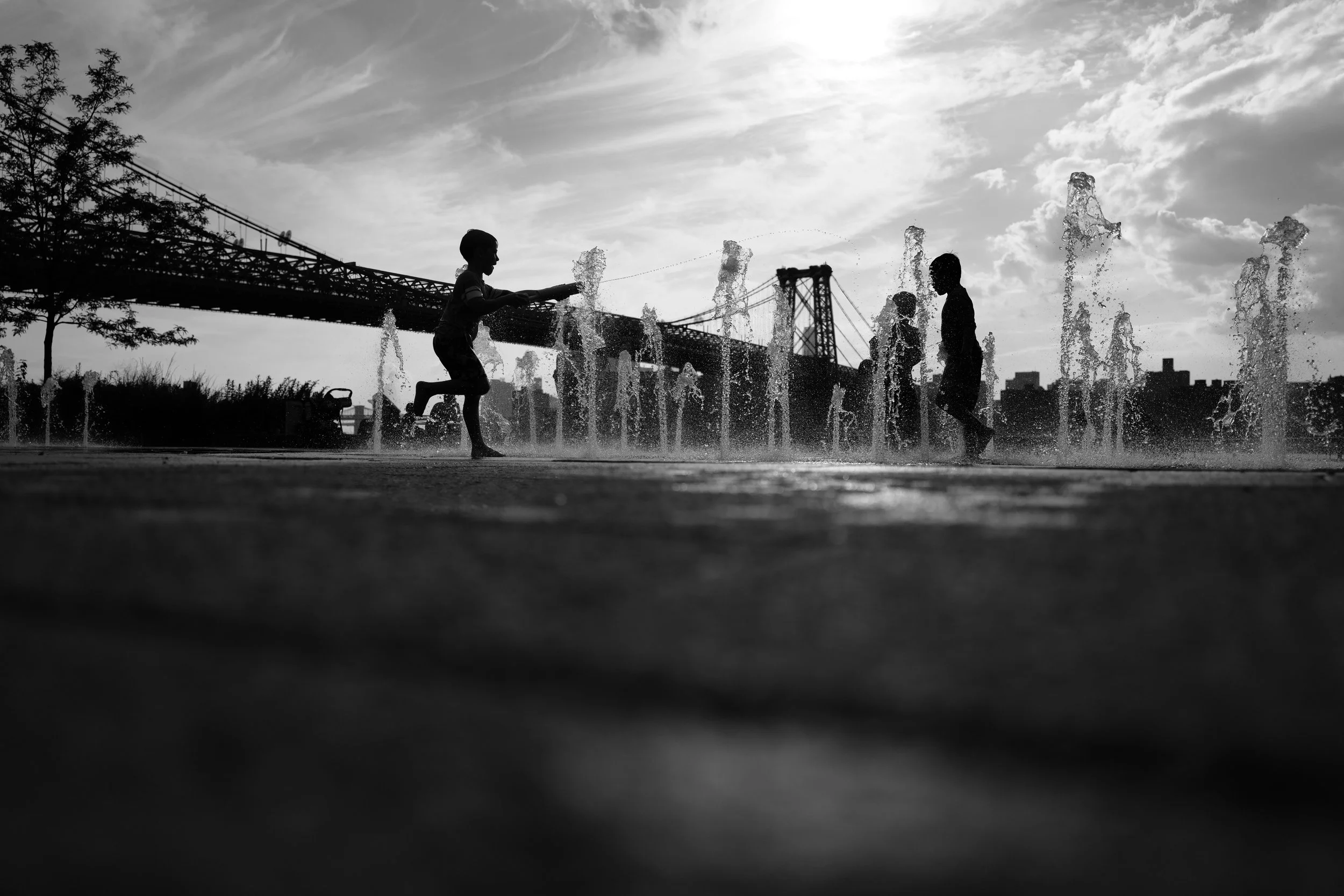 Children playing in a splash pad or water fountain at sunset with a bridge and city skyline in the background.