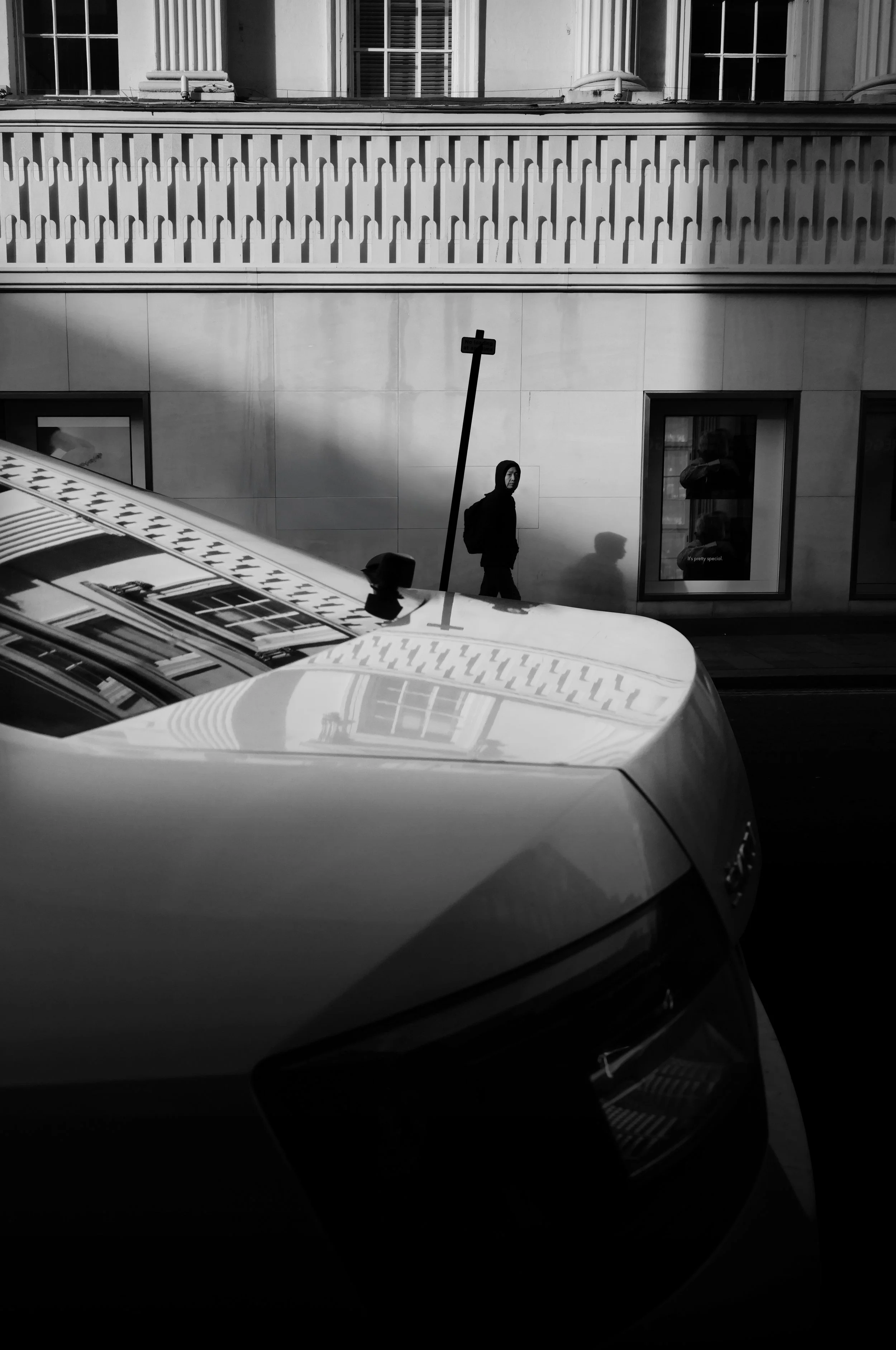 Black and white photo of a city street scene with a parked car in the foreground, a pedestrian in a hoodie walking, a street sign on a pole, and a building with columns and windows in the background.