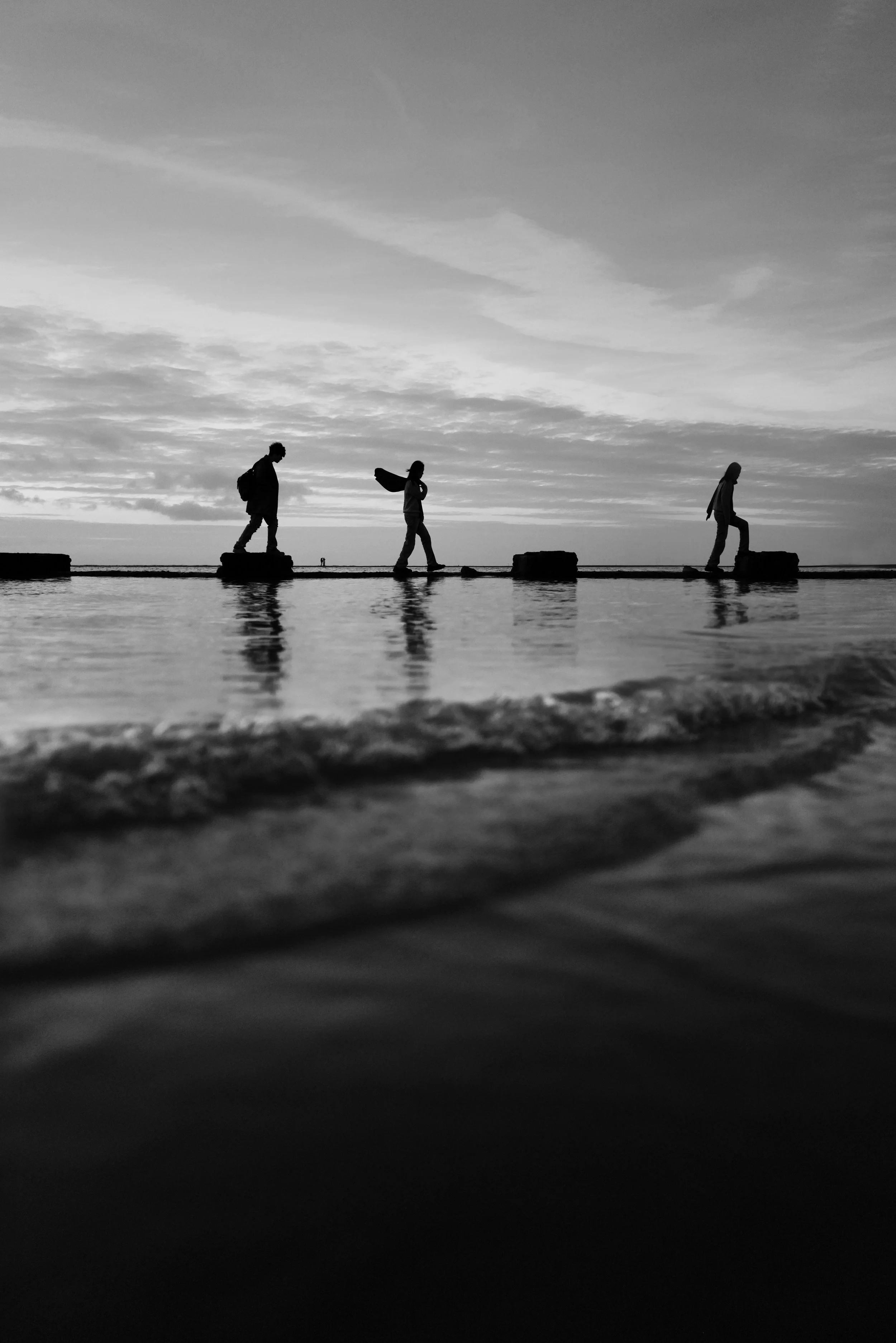 Silhouettes of three people walking on a pier by the water at sunset, reflected on the water's surface, black and white photo.