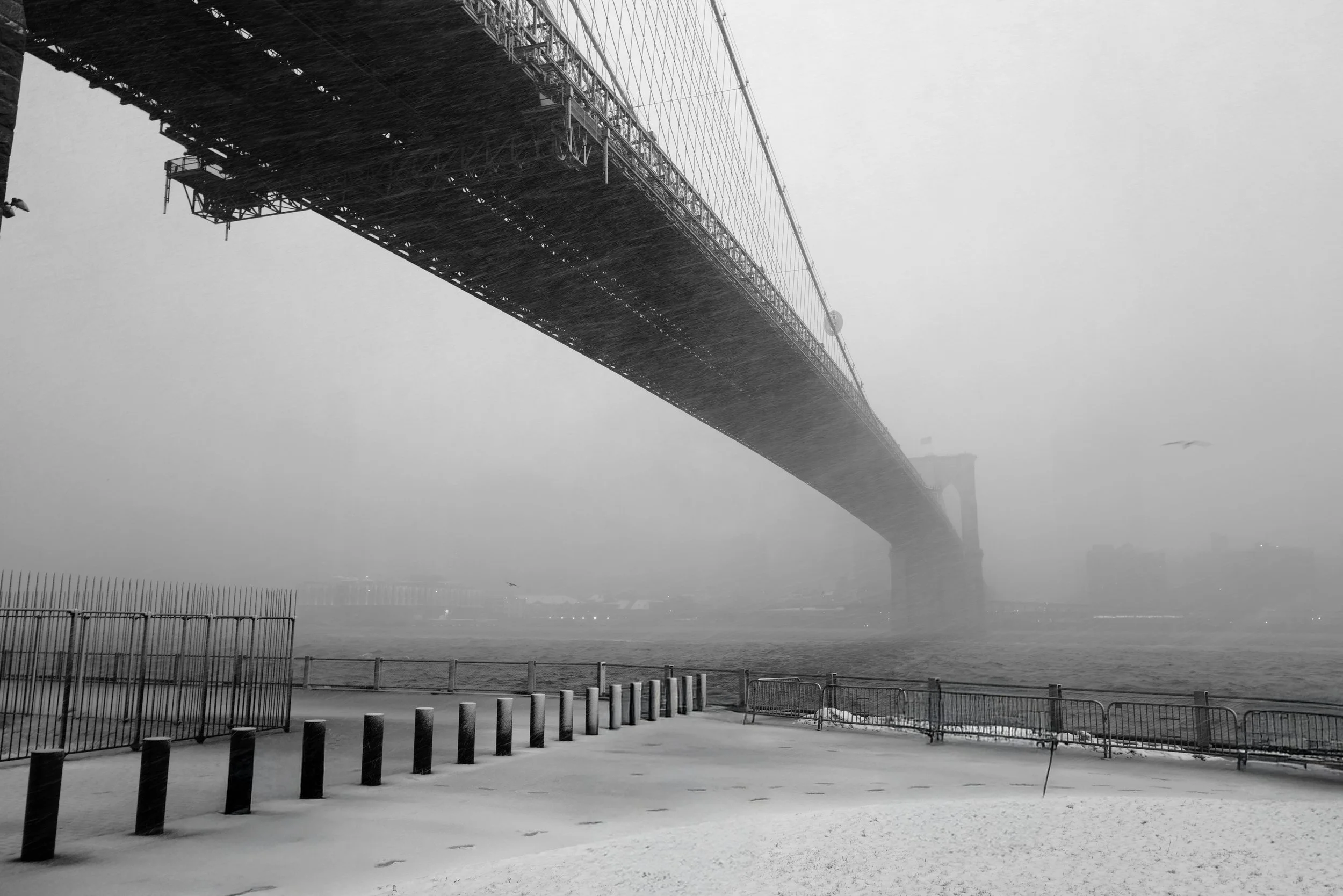 A black and white photo of a large suspension bridge over a body of water with snow in the foreground and a foggy, overcast sky.