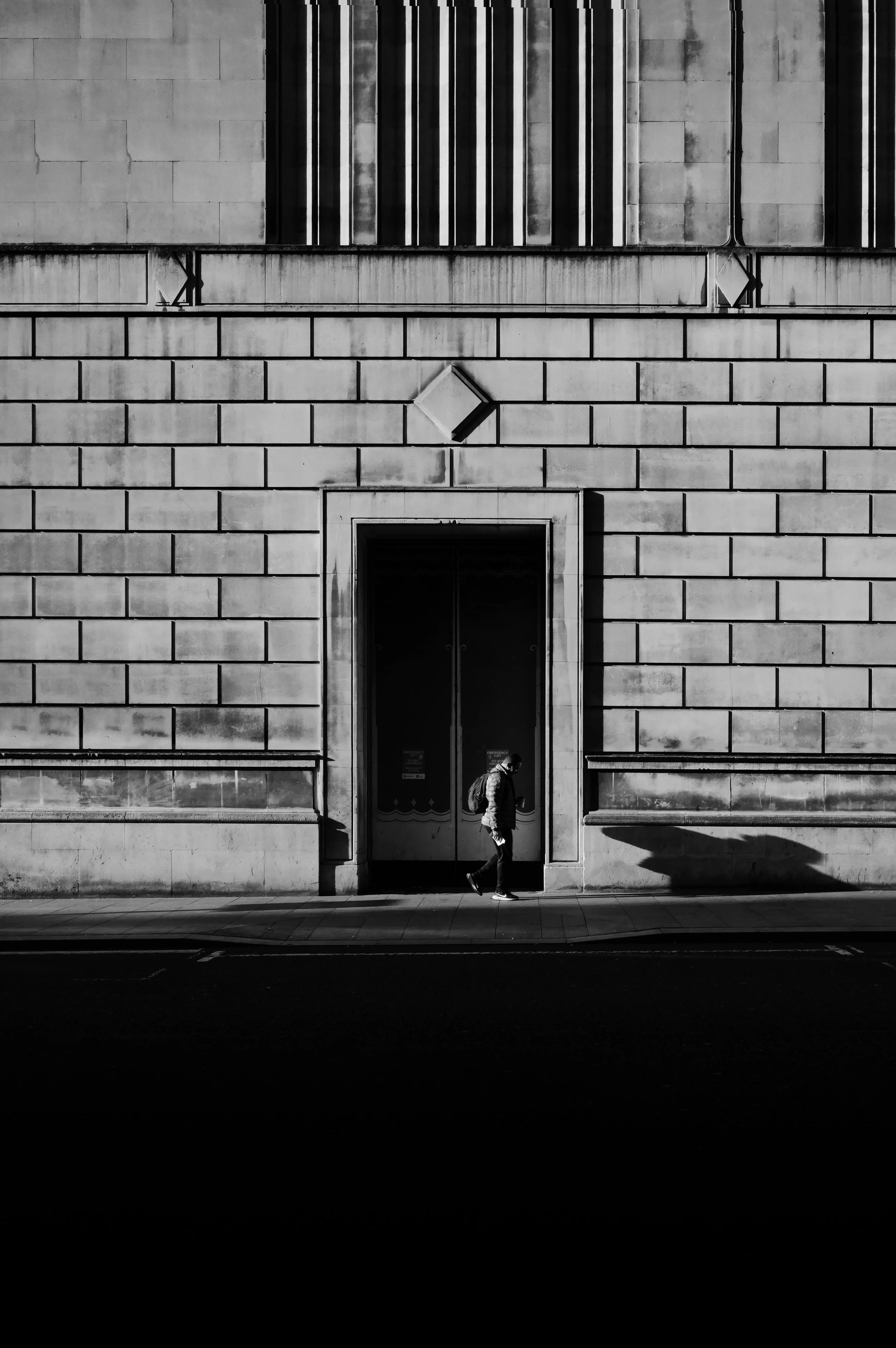 A person walking past a large, historic building with a brick facade, tall doors, and vertical striped windows in black and white.