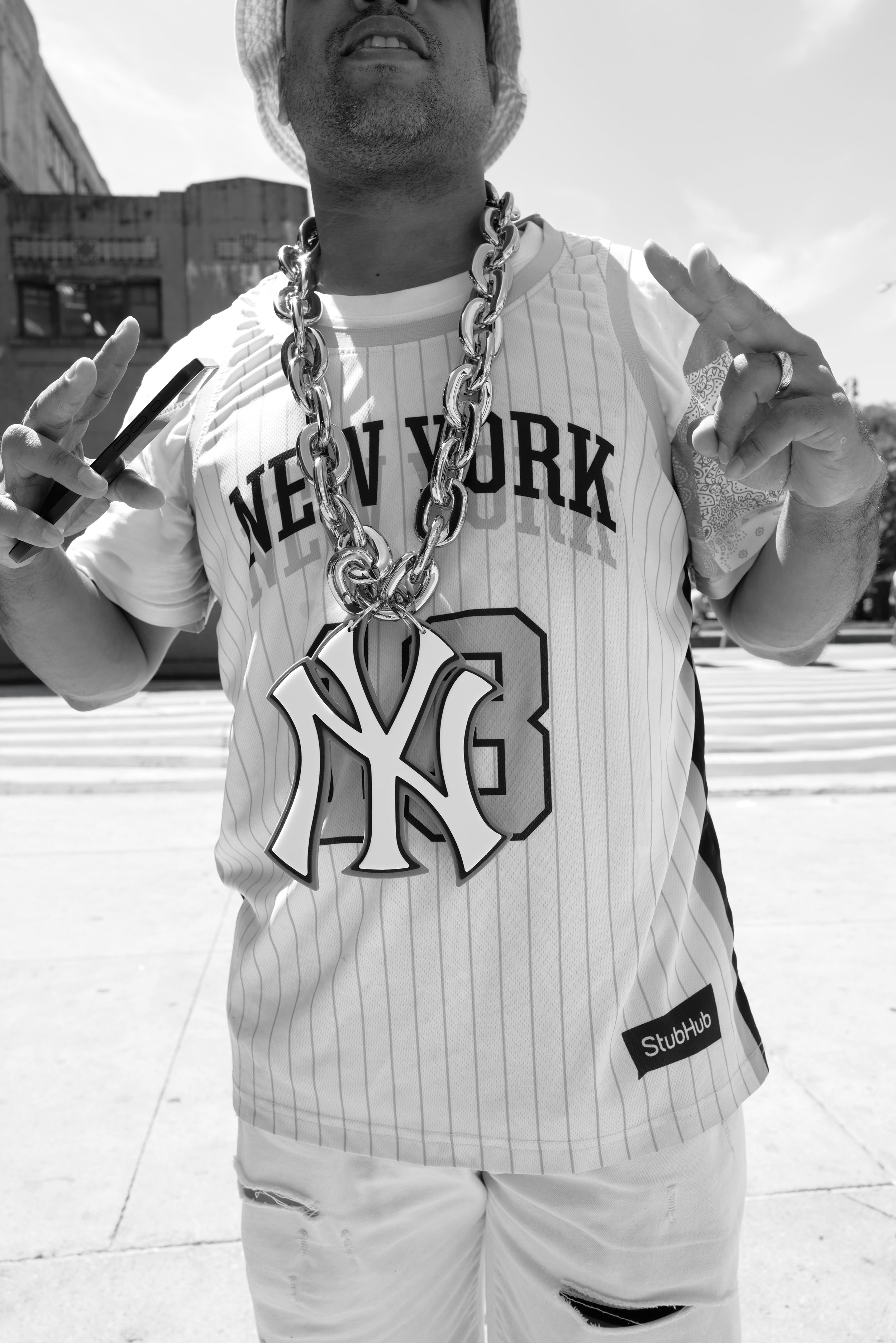 A man wearing a New York Yankees baseball jersey, large chain necklace with Yankees logo, hat, and holding a phone, standing outdoors.