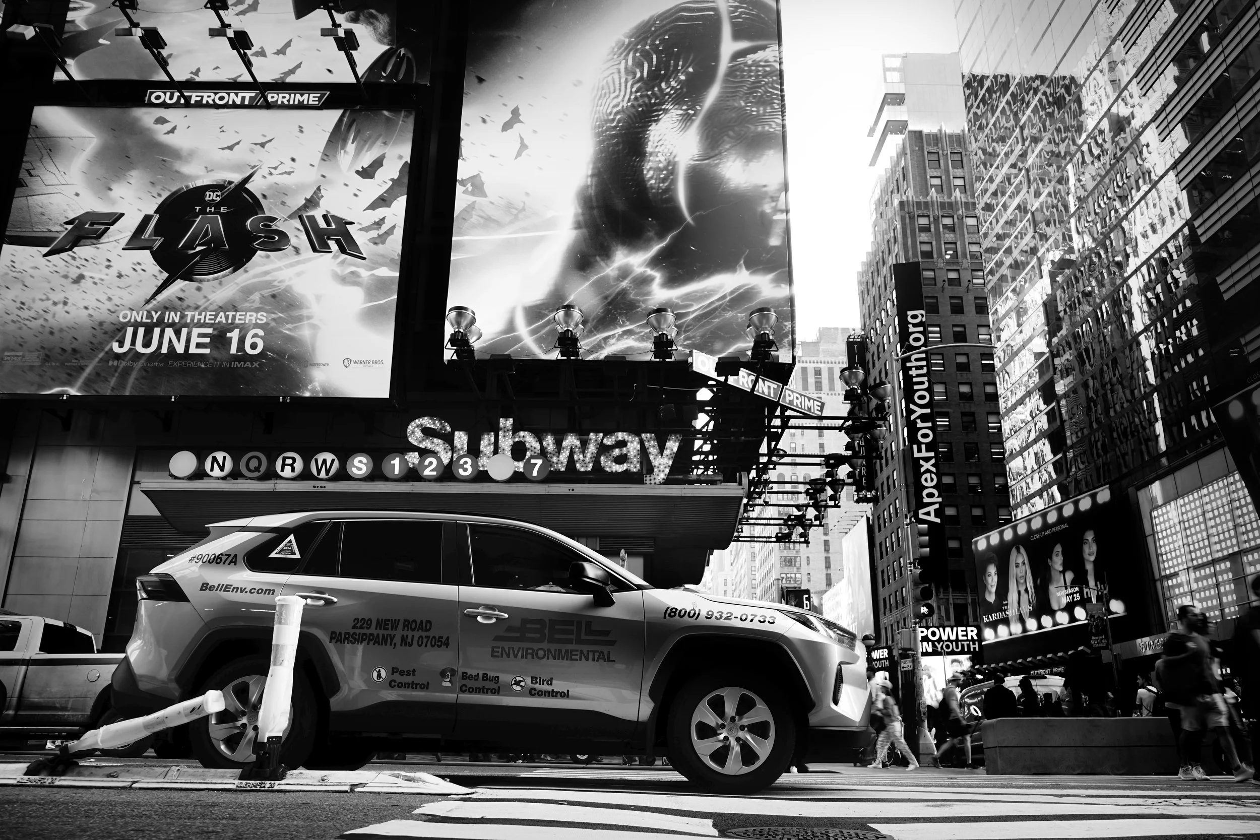 Black and white street scene featuring a subway entrance, a parked environmental service vehicle, illuminated billboards, tall buildings, and pedestrians in Times Square, New York City.