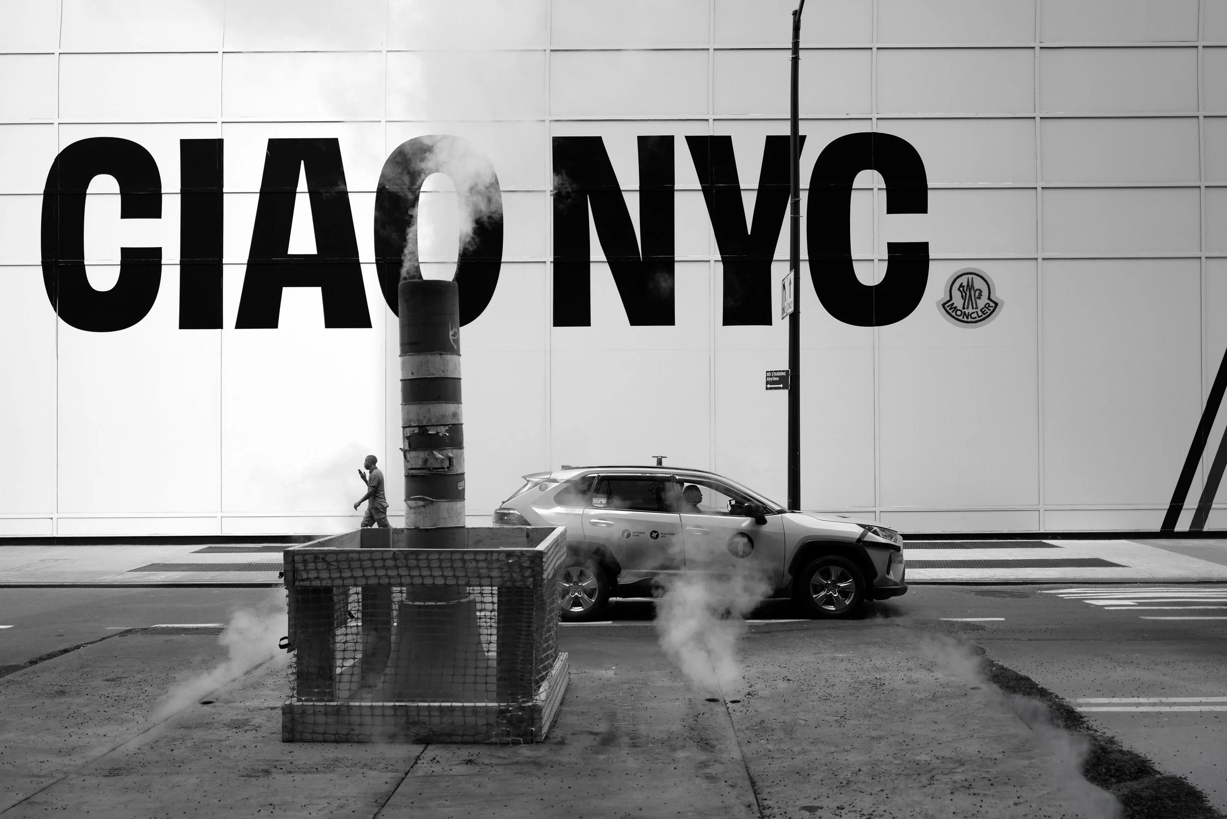 Black and white street scene with a large wall in the background displaying the text 'CIAO NYC'. In the foreground, there is a car parked on the street, a street lamp emitting smoke or steam, and a person walking on the sidewalk while looking at their phone.