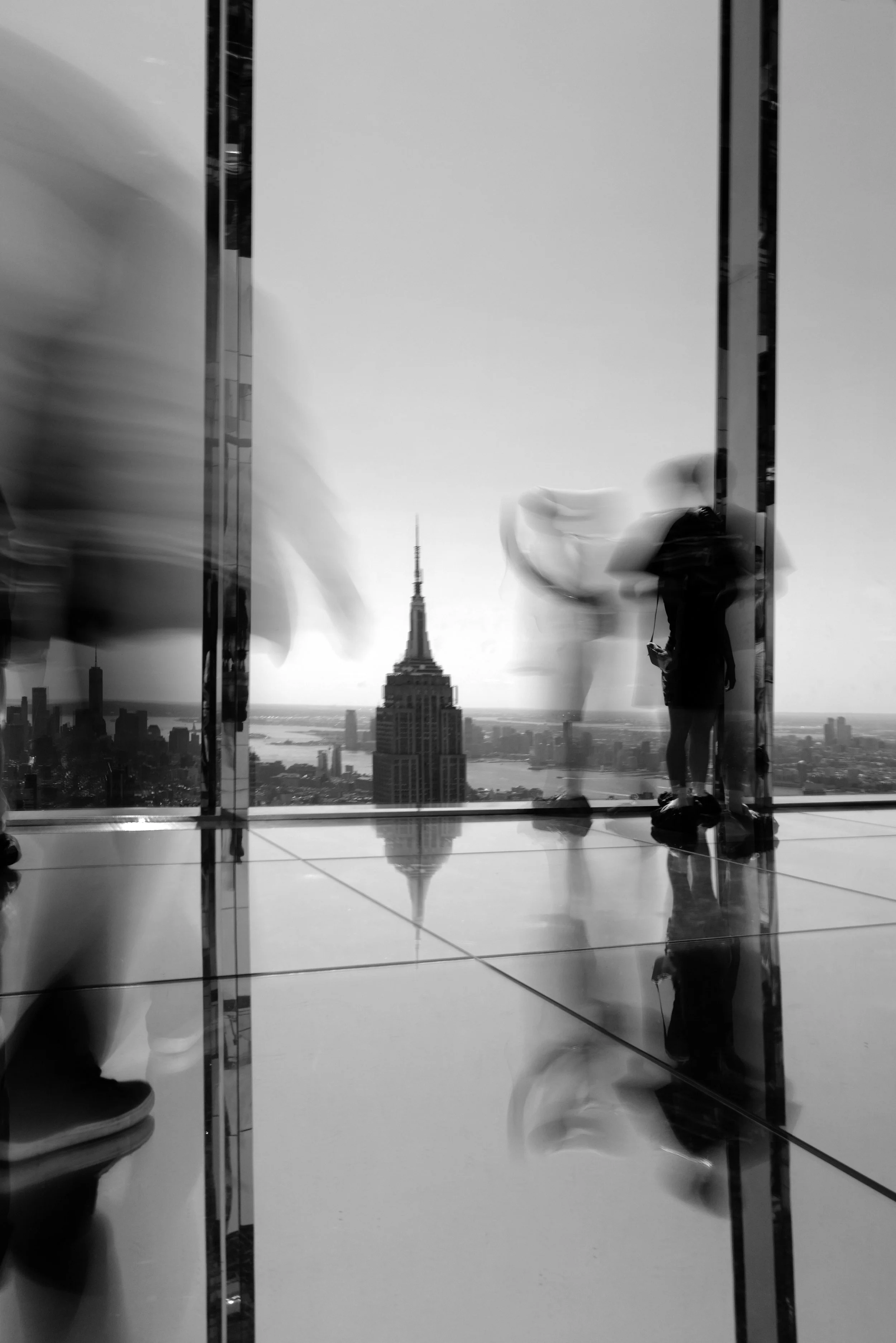 View from inside a modern skyscraper with glass walls, showing blurred reflections of people walking, and the Empire State Building in the distance in New York City.