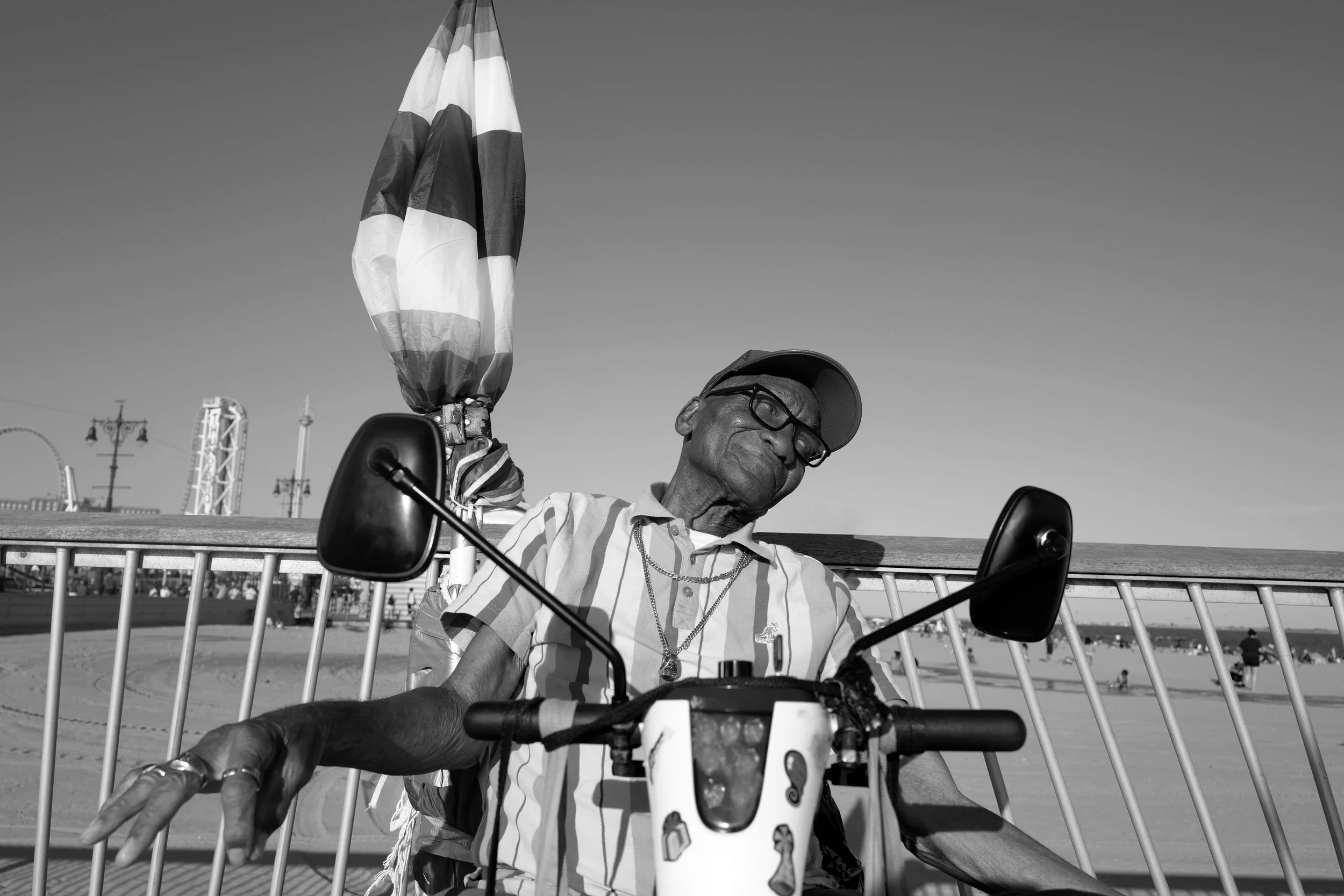 An elderly man wearing a cap, glasses, a striped short-sleeve shirt, and jewelry, sitting on a motorized scooter at the beach with amusement park rides visible in the background, in black and white.