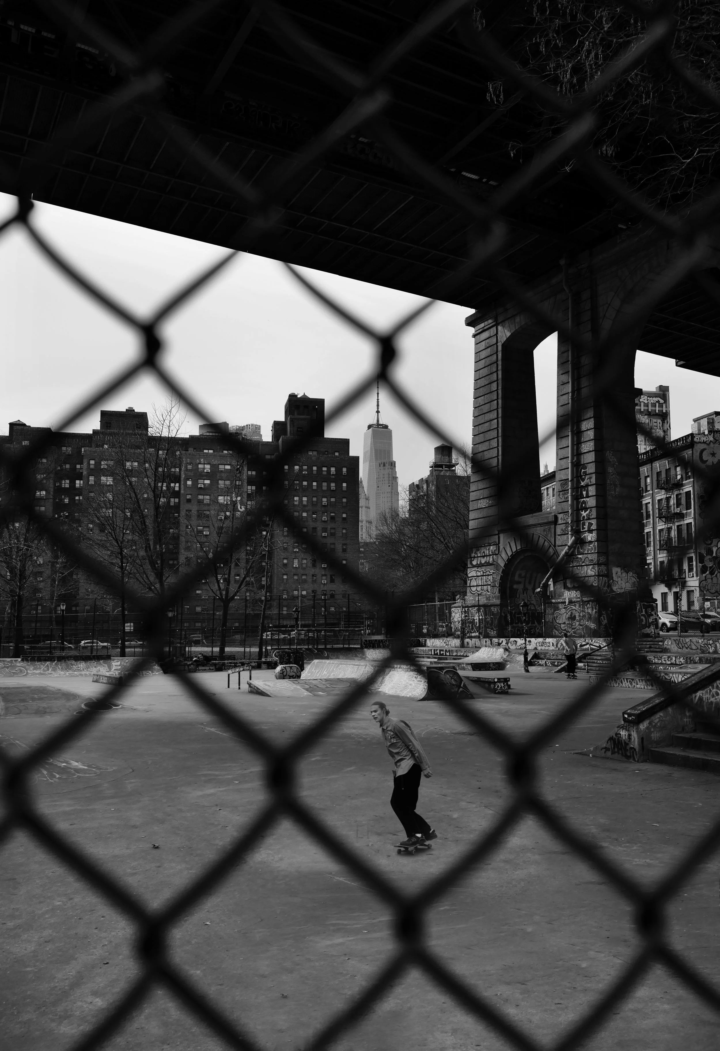 Skateboarder skating at an urban skate park with city buildings and a bridge overhead, viewed through a chain-link fence in black and white