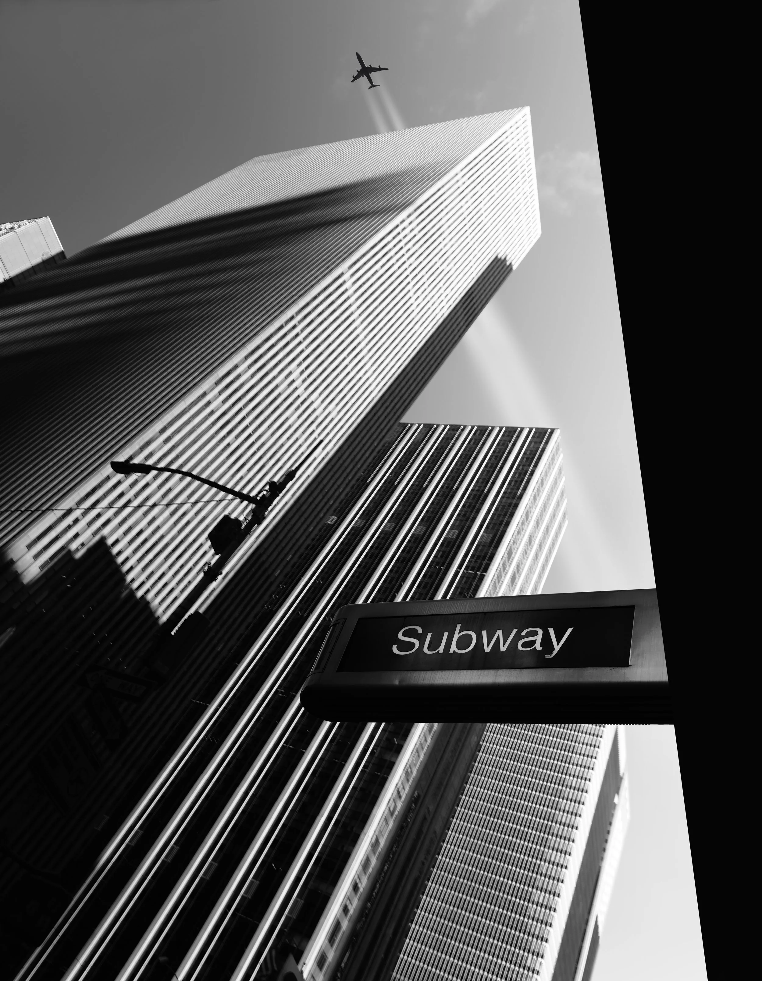 Black and white photo of skyscrapers in New York City with the Twin Towers and a plane flying overhead, viewed from below near a subway entrance.