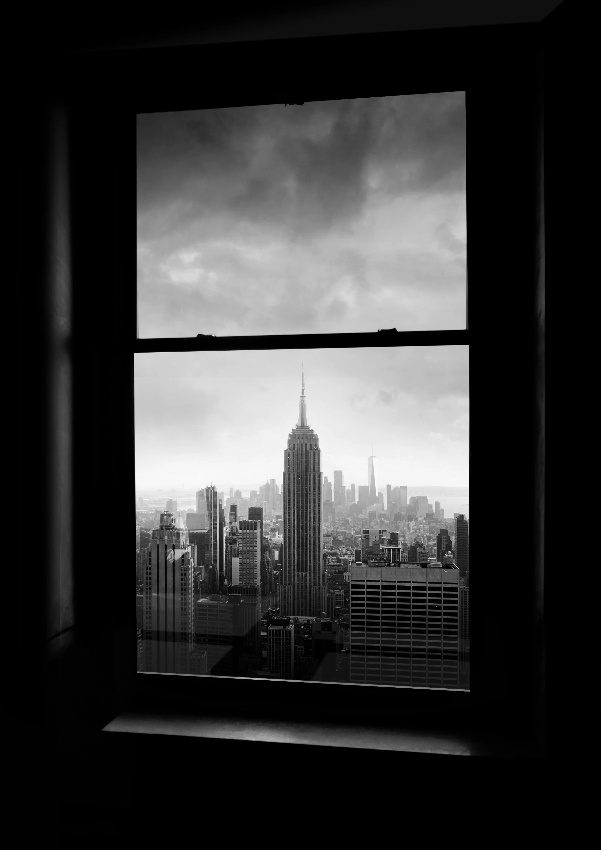 View of the New York City skyline through a window, with the Empire State Building prominently in the center and other skyscrapers in the background, in black and white.