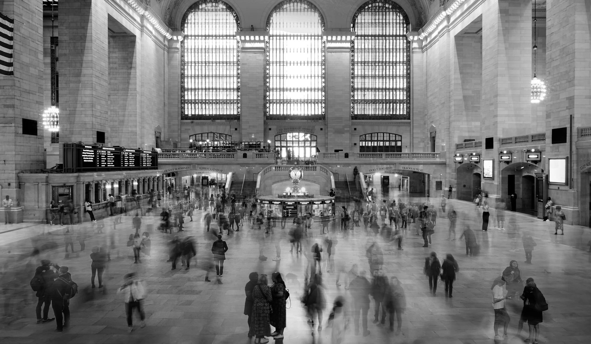 Interior of a grand train station with high arched windows, a central clock, and many people walking on the marble floor.