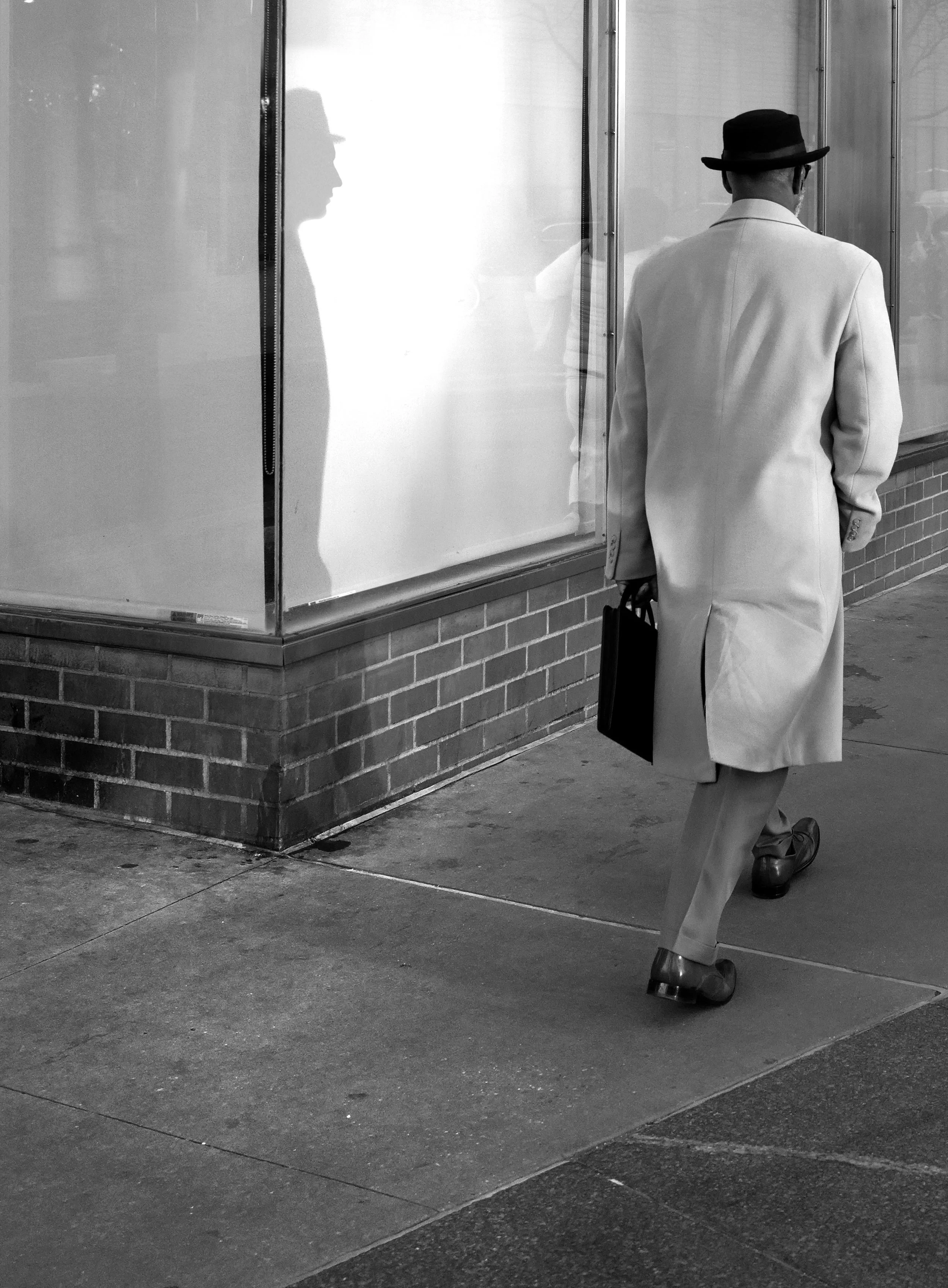 A man in a light-colored overcoat and hat walking past a glass window with a reflection of himself, carrying a briefcase.