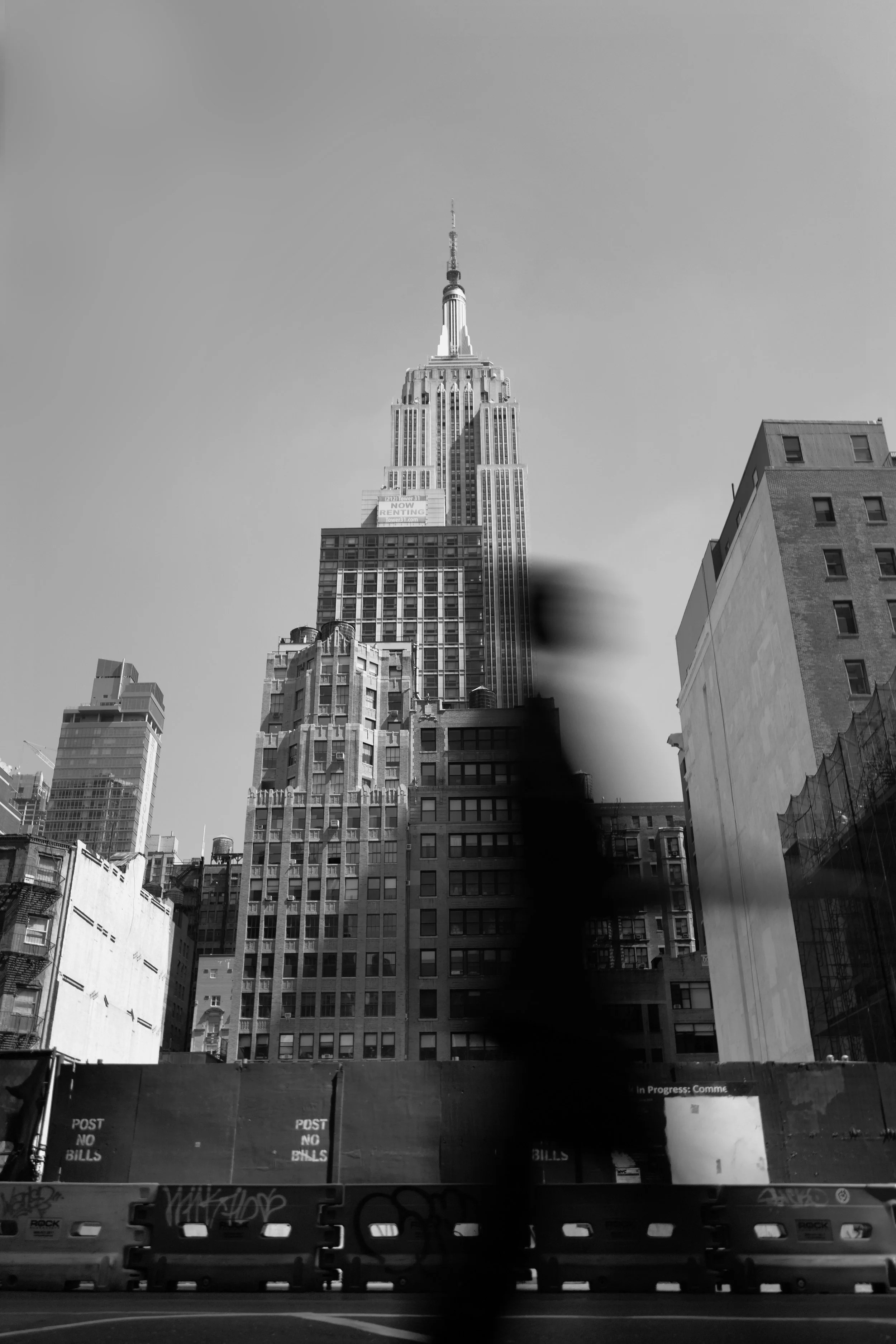 Black and white photo of the Empire State Building in New York City, surrounded by other tall buildings, seen from street level.