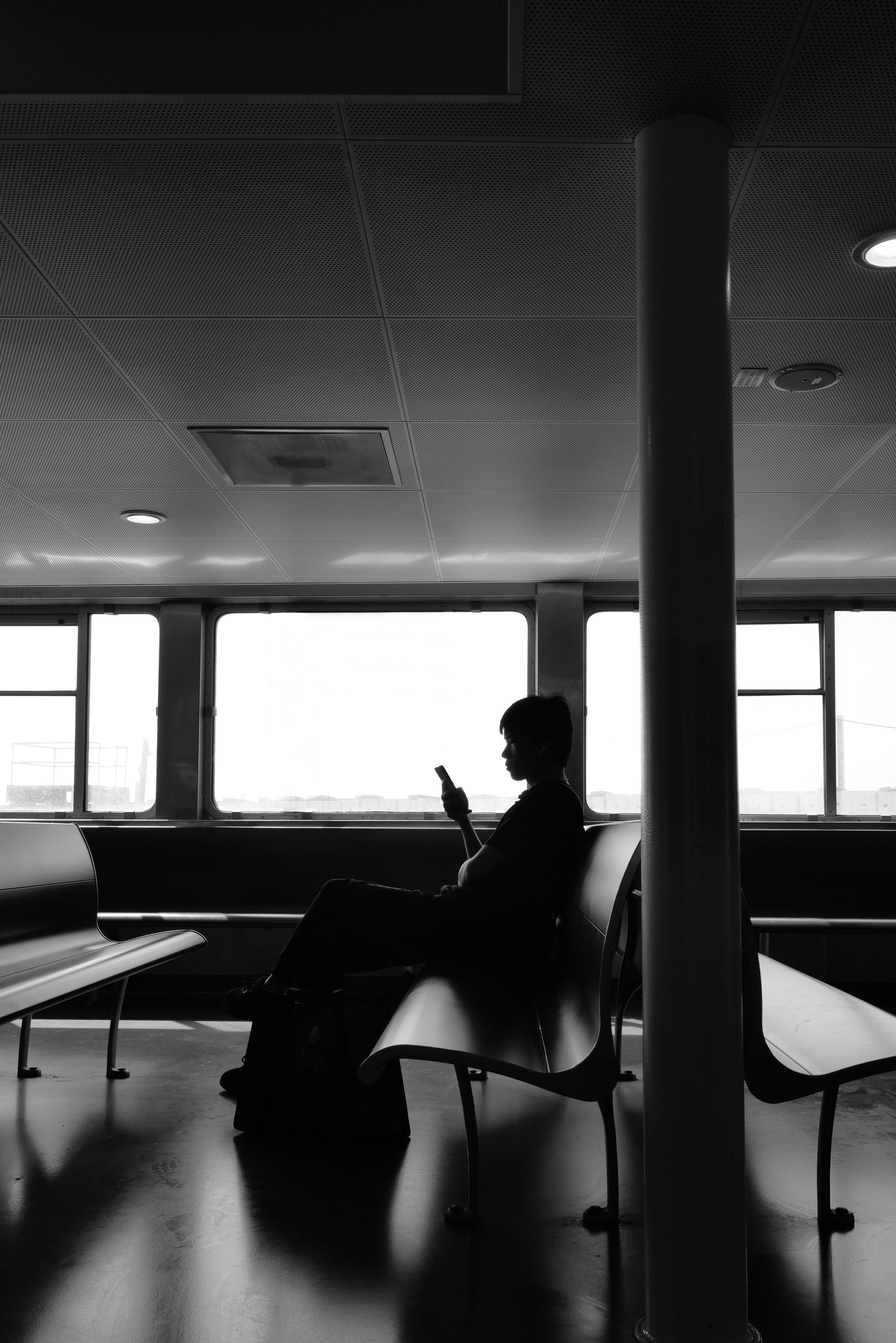 Silhouette of a person sitting alone on a bench, looking at their phone, inside a waiting area with large windows showing an outdoor view.