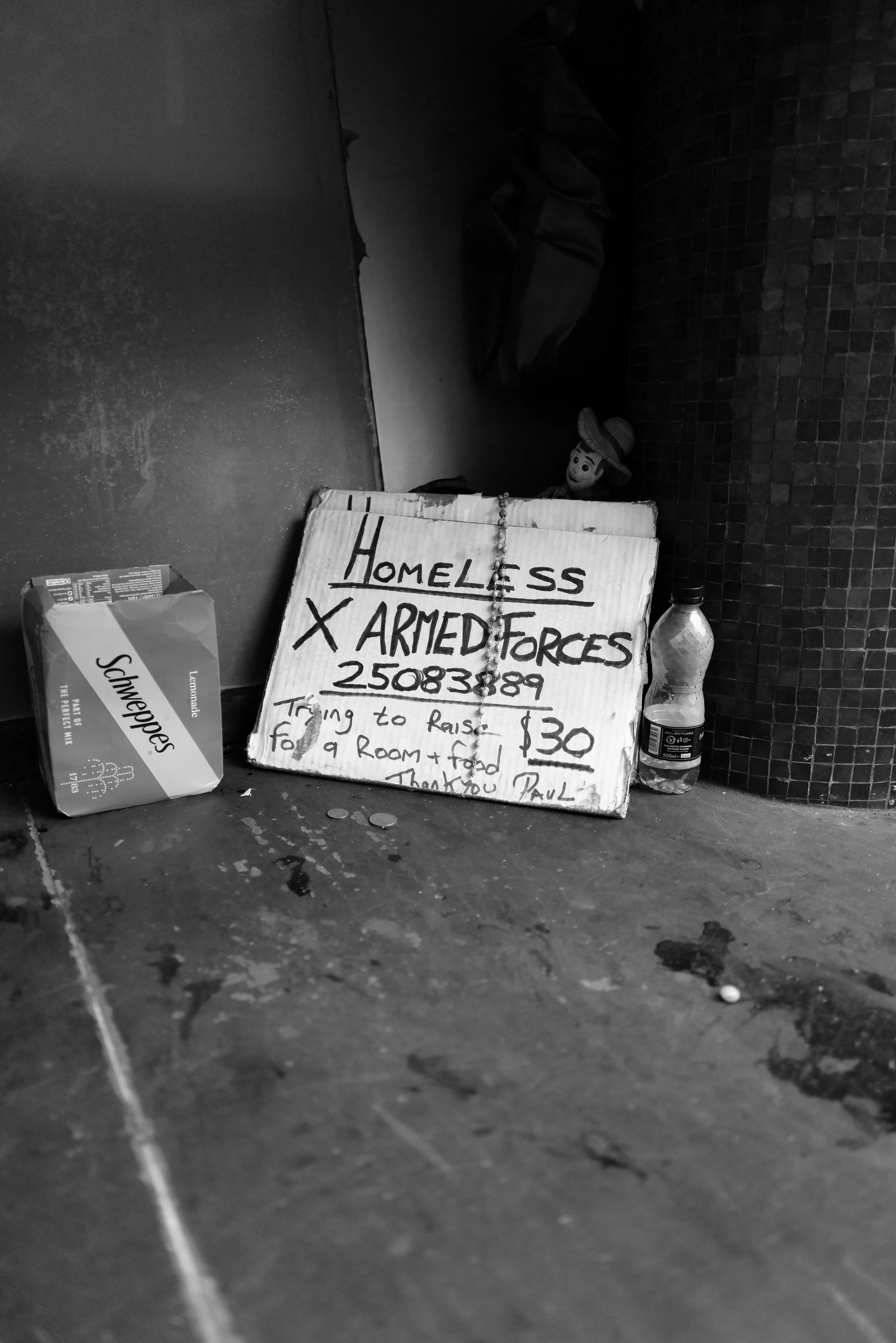 A handwritten sign on cardboard offering help for homeless people, promoting fundraising for a room and food, with a phone number and pricing details. Items include a box of Schweppes Tonic Water, a plastic water bottle, and a small doll or figurine in the background, placed on the ground next to a tiled pillar.