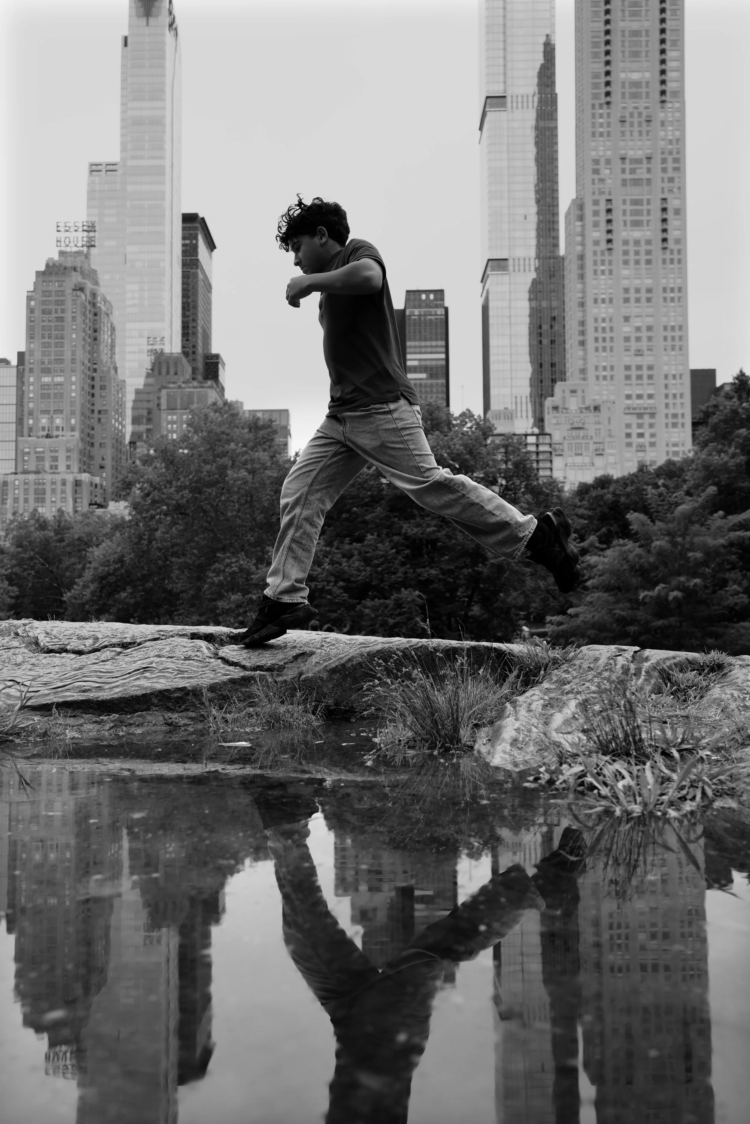 A person is running on a rock near a small pond in a park with tall skyscrapers in the background, black and white photography.
