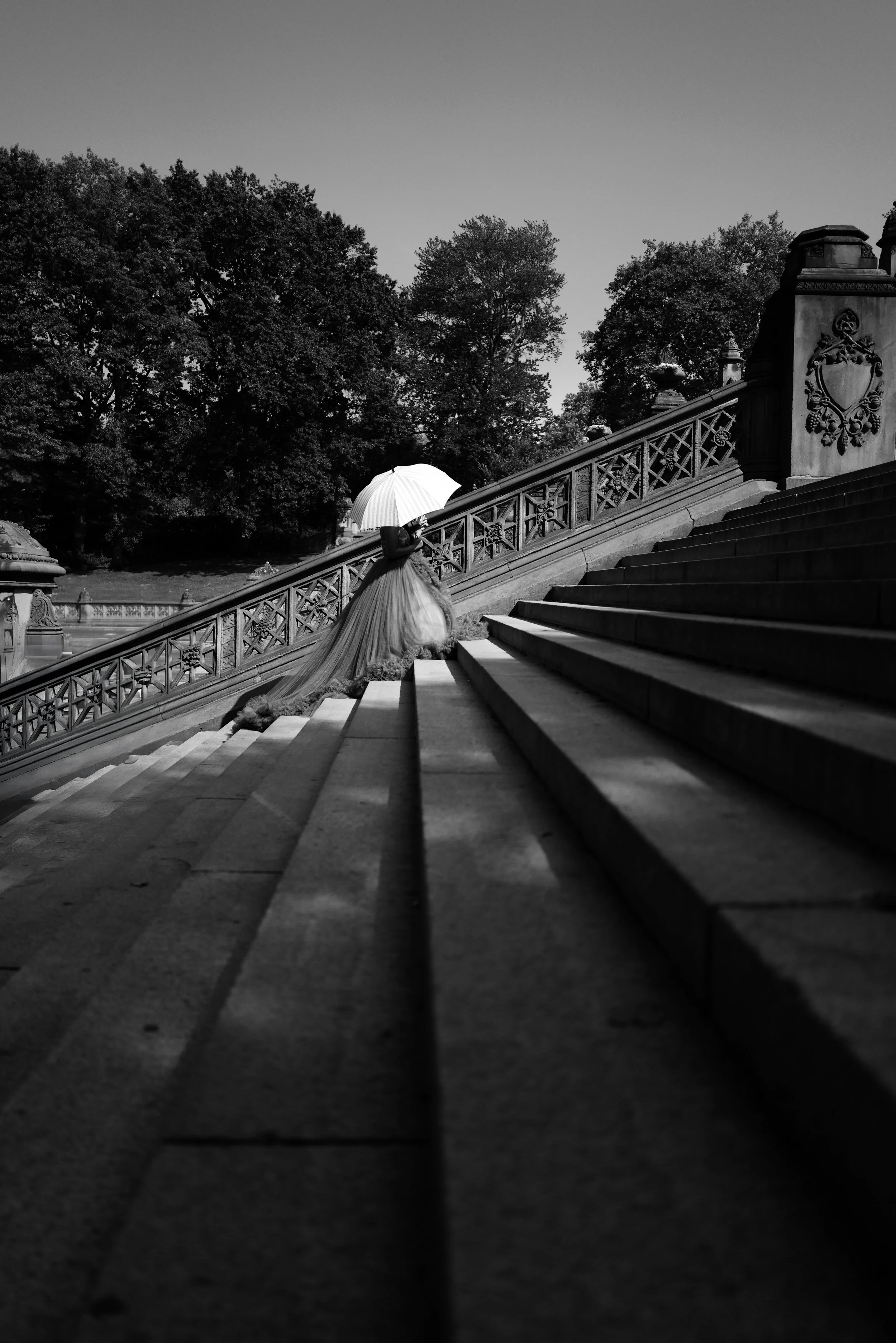 A woman in a flowing dress holding an umbrella ascending outdoor stone stairs with ornate railings, trees in background, black and white photo.