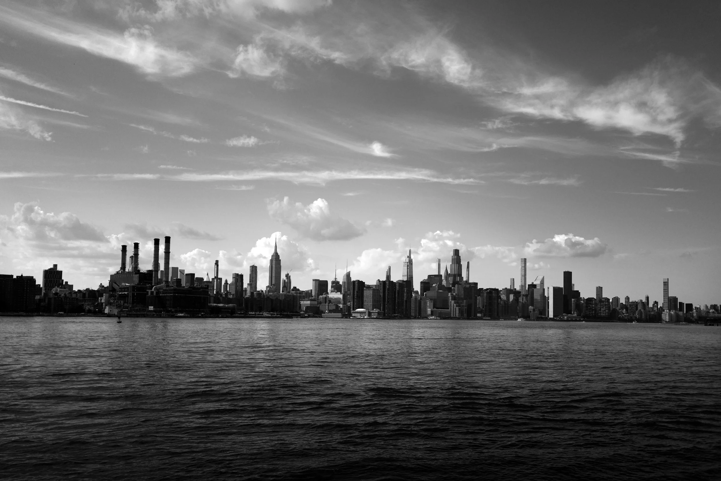 Black and white photo of the New York City skyline seen across the water, including the Empire State Building and other tall skyscrapers, under a partly cloudy sky.