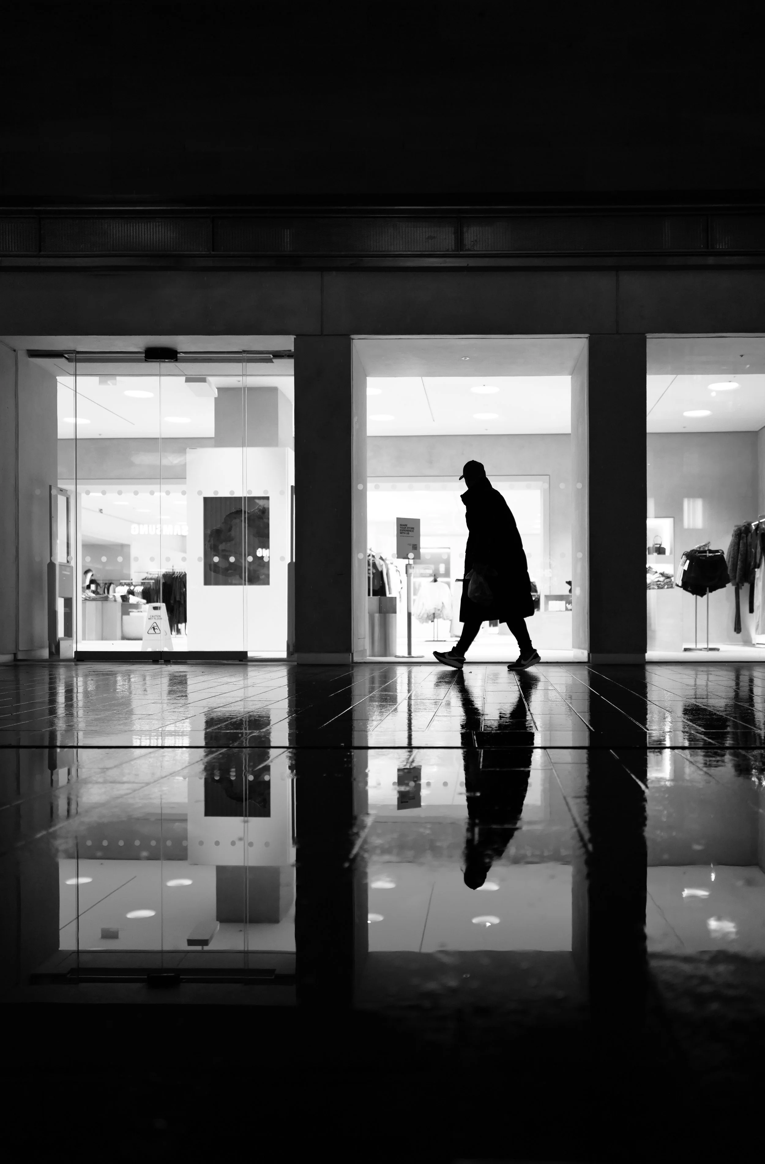 A silhouette of a person walking past a brightly lit store with mannequins and clothing displays, reflected on a wet floor in black and white.