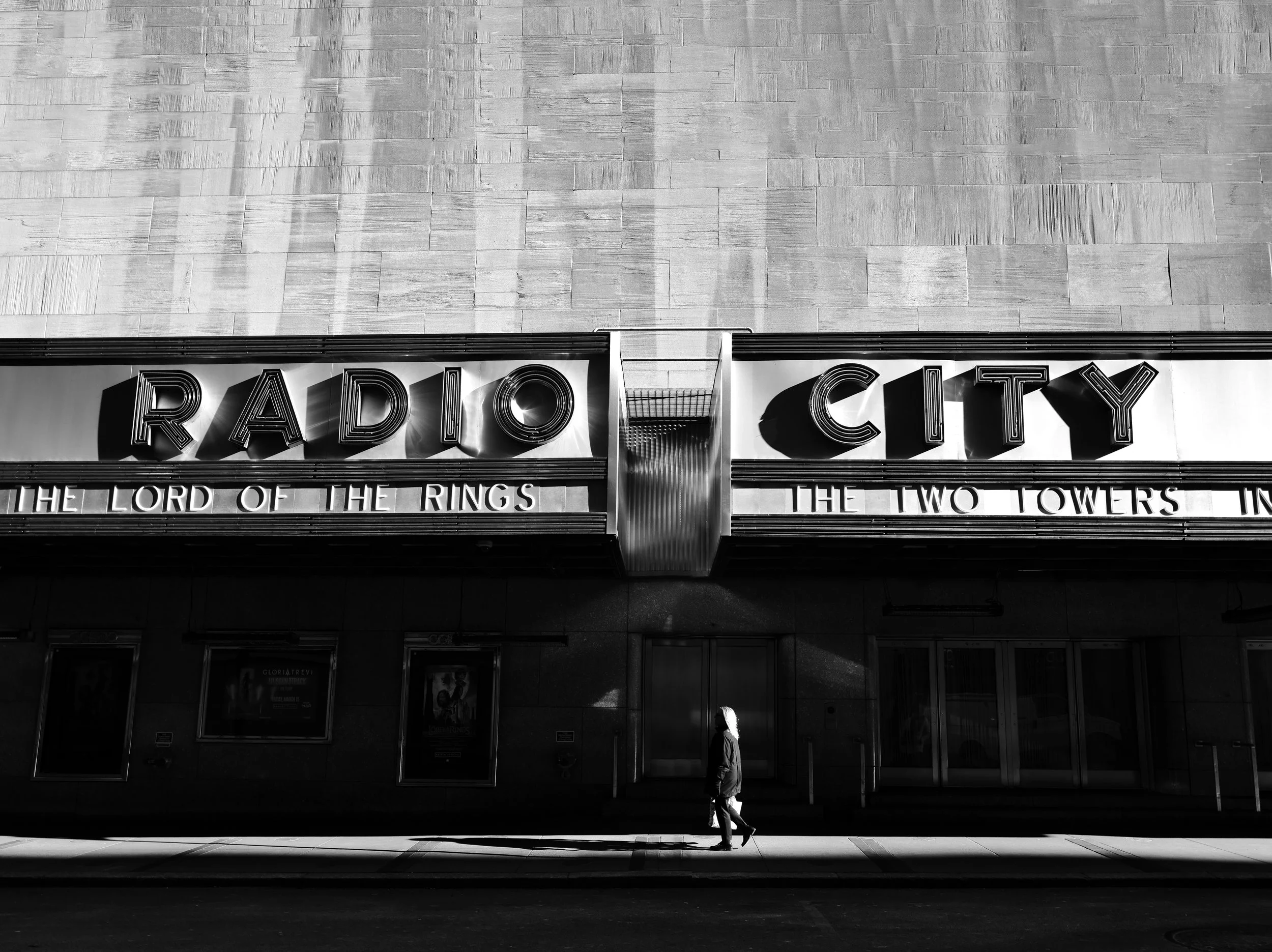 A black and white photo of the exterior of Radio City Music Hall with a person walking past in the foreground