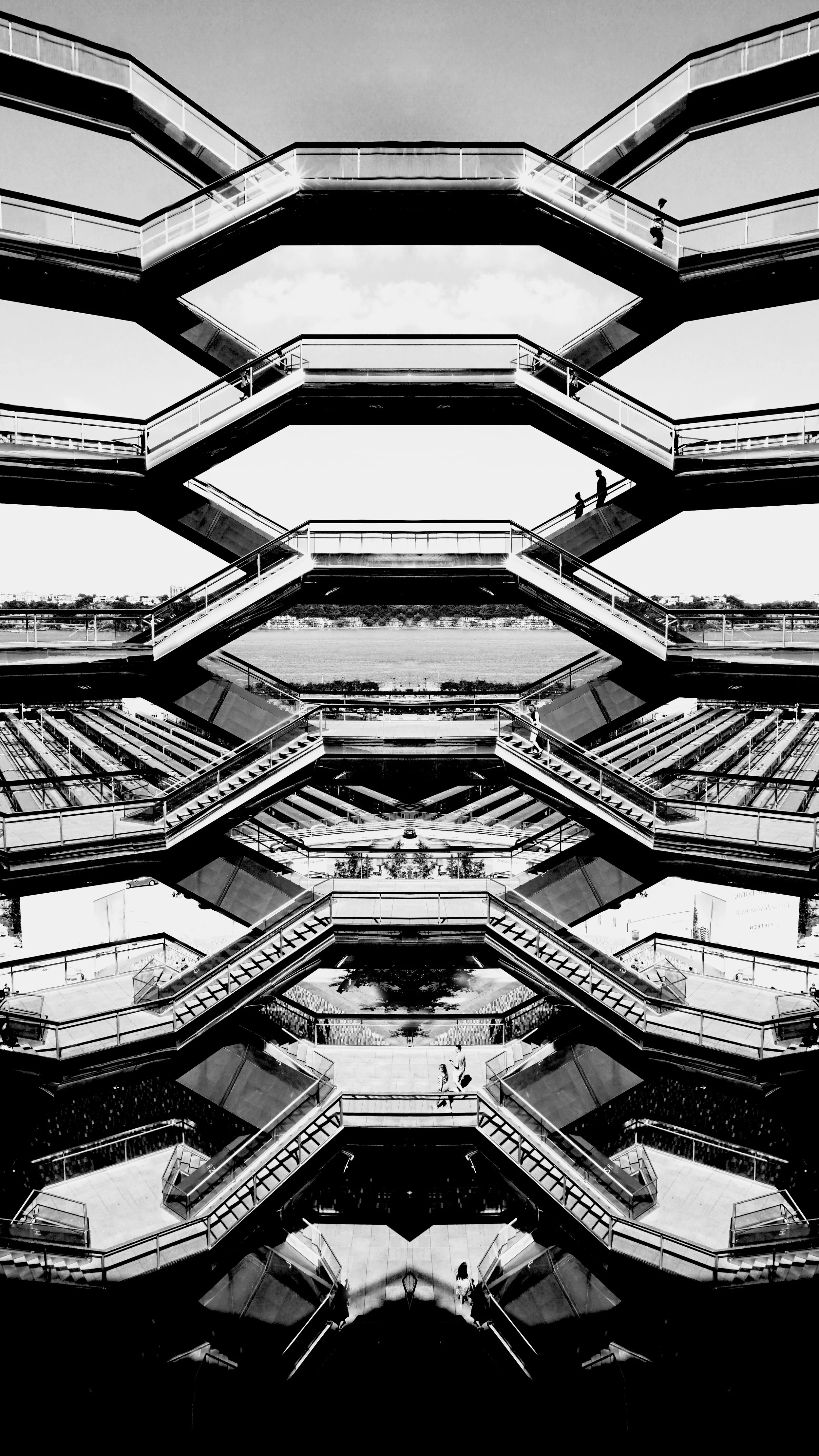 Black-and-white photo of a modern, multi-level outdoor staircase structure with glass railings, featuring geometric shapes and symmetry, with a view of water and a distant landscape in the background.
