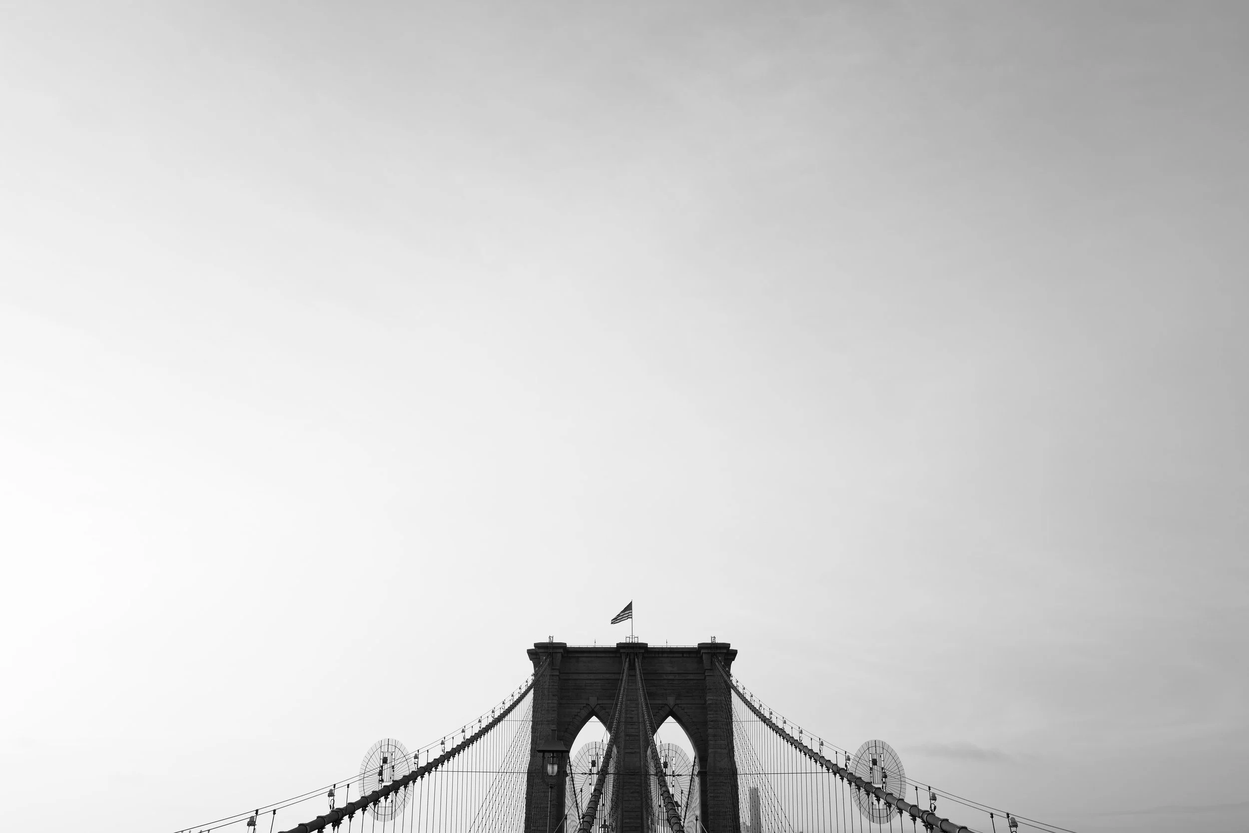 Black and white photo of Brooklyn Bridge with an American flag on top, suspension cables, and city skyline in the background under a cloudy sky.