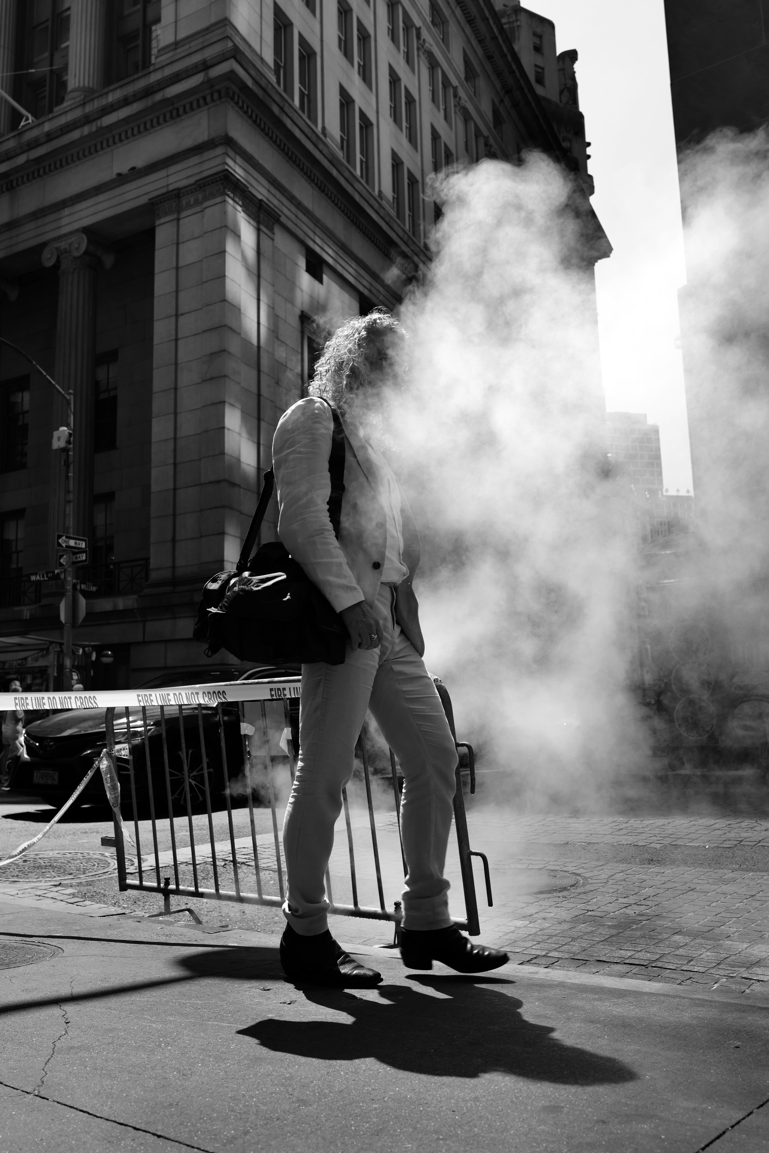 A woman in business attire walking past a smoke-filled street scene, with police barricades and tall buildings in the background, in black and white.