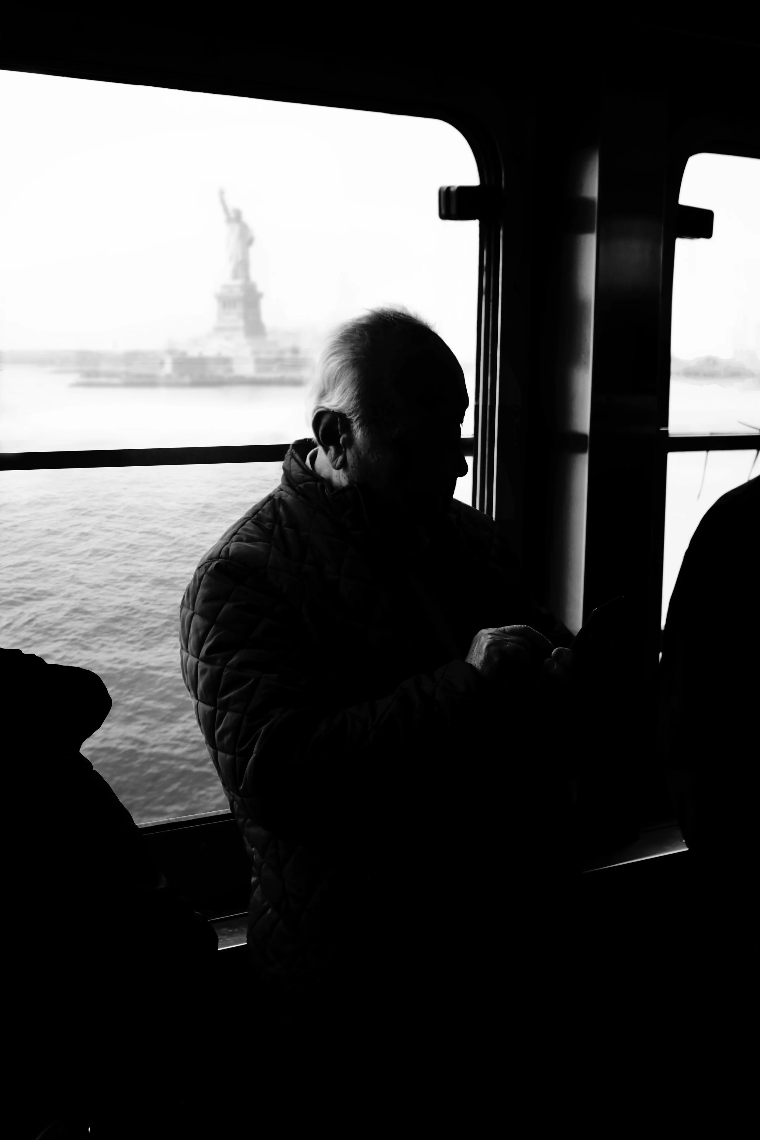 A silhouette of an elderly man sitting inside a boat, with the Statue of Liberty visible in the background through the window.