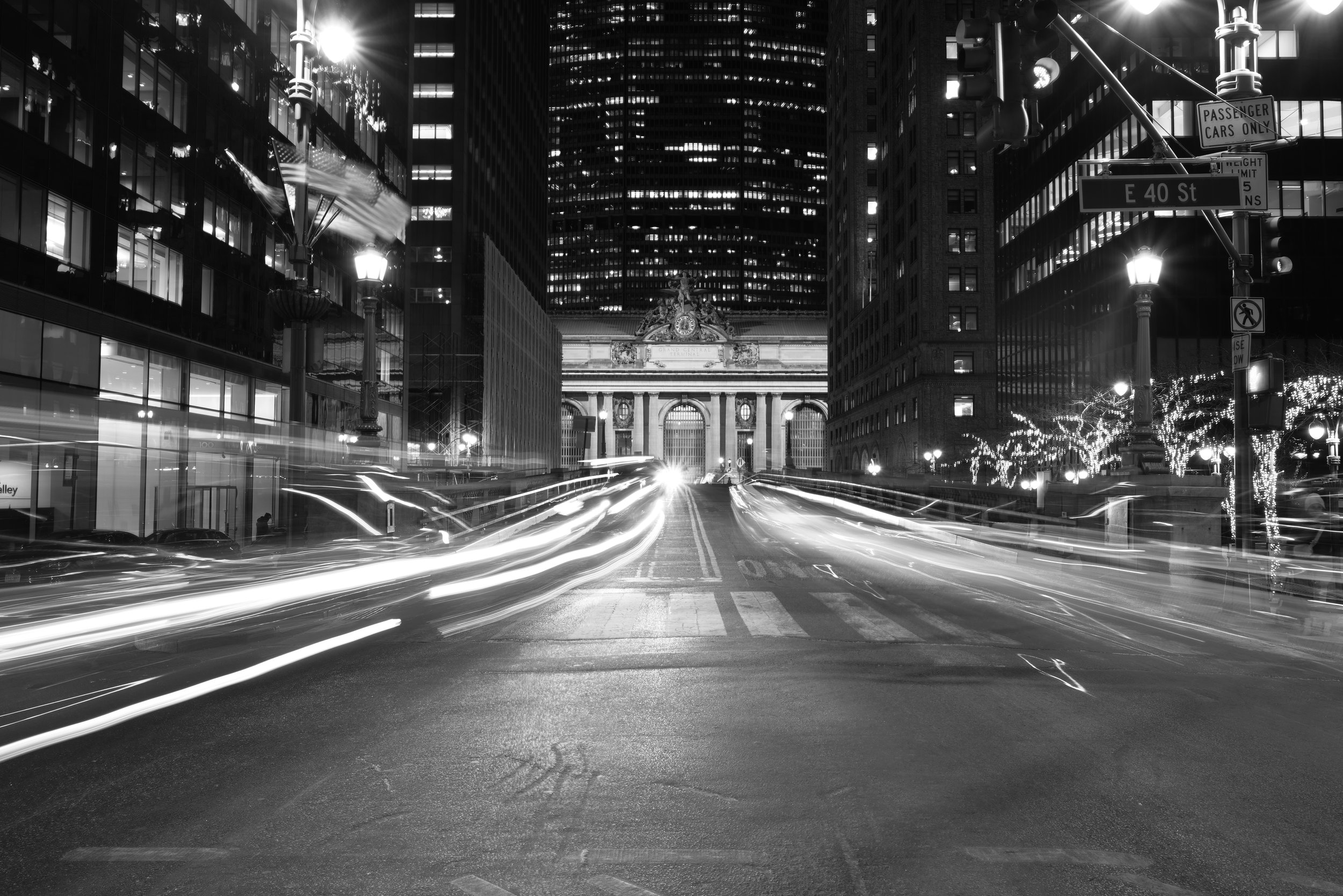 Black and white nighttime city street with streaks of car lights, tall skyscrapers, and historic building at the end of the street.