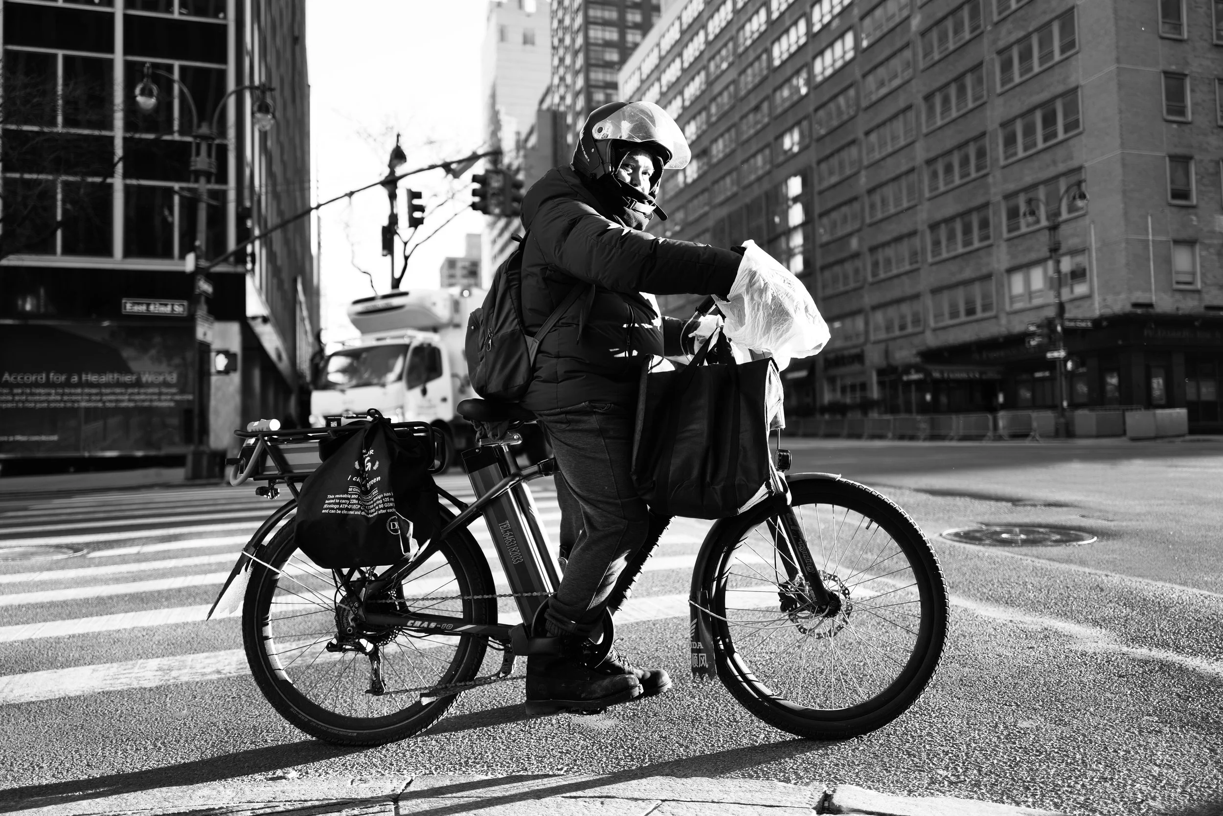 A person wearing a helmet and a jacket riding a bicycle across an urban street with tall buildings, some carrying bags on the bike, black and white