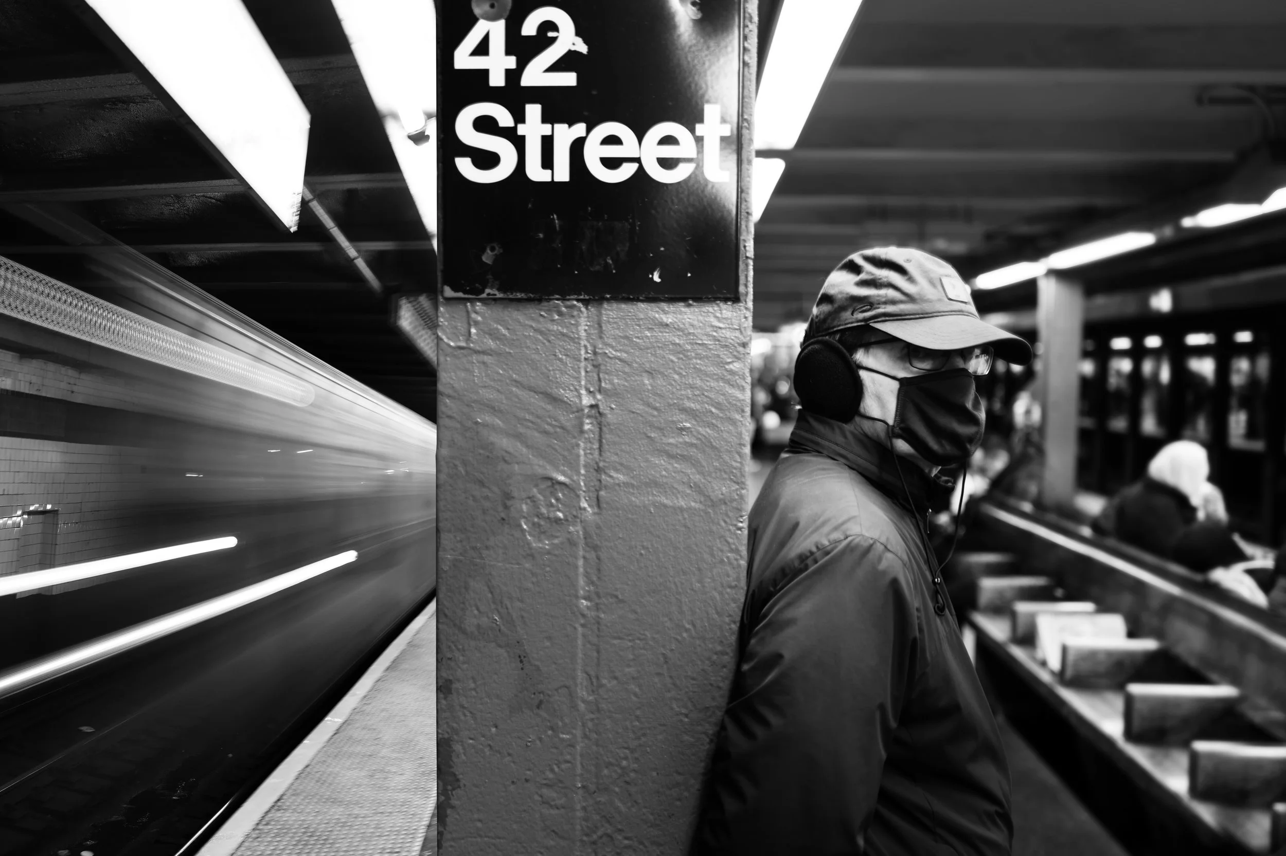 A man wearing a cap, face mask, headphones, and glasses standing on a subway platform with a blurred train passing by in the background, at 42nd Street.