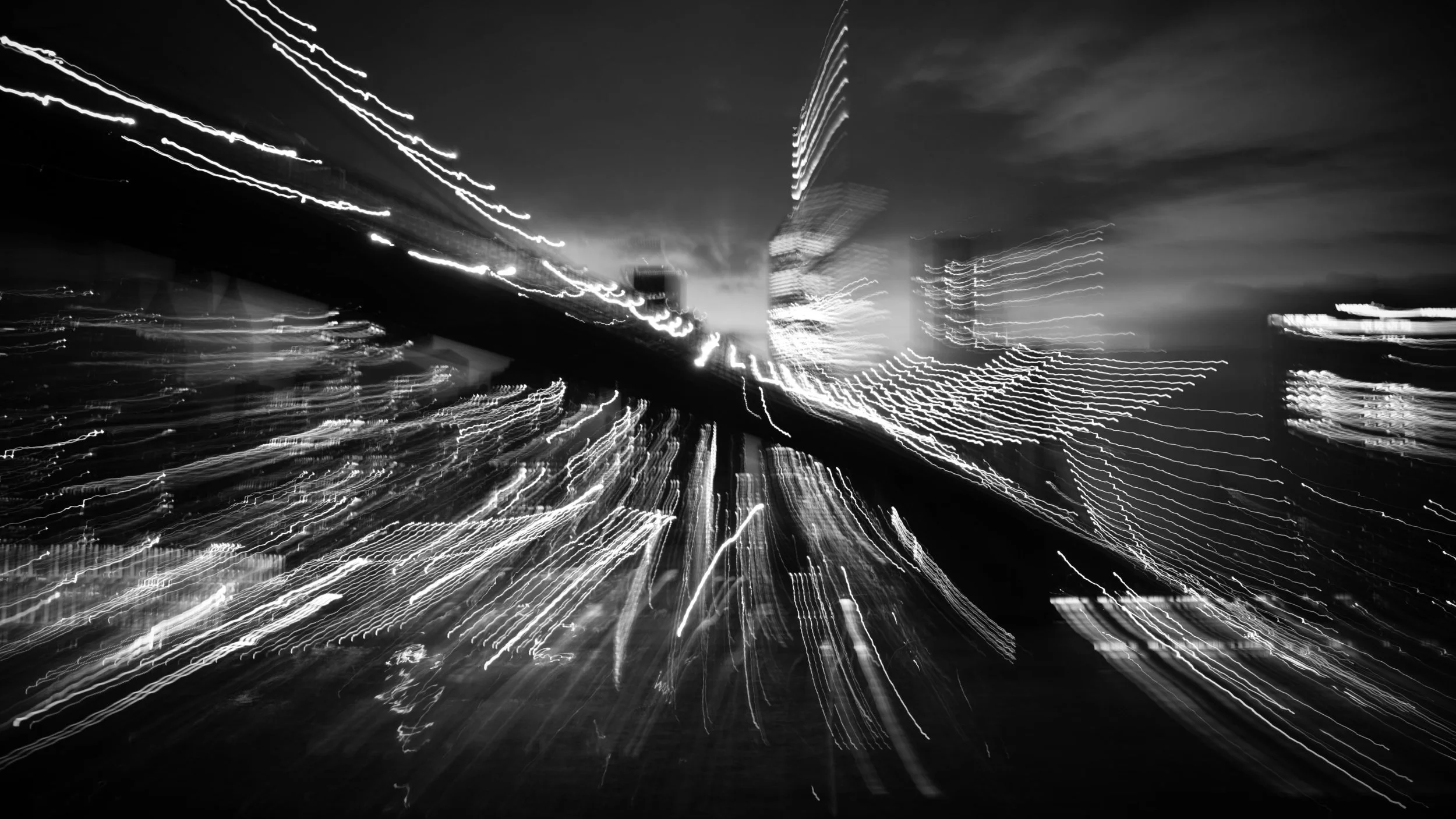 Blurred black and white photo of a cityscape at night with streaks of lights from moving vehicles and buildings, creating a dynamic abstract effect.