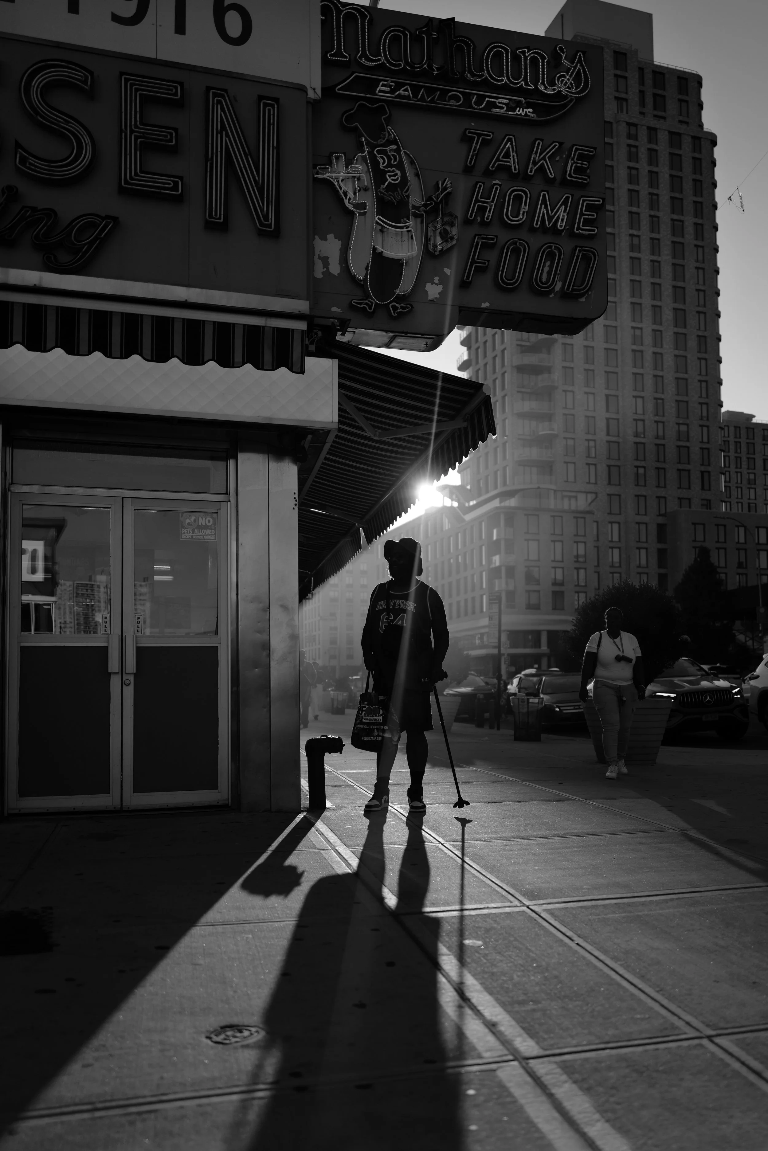 A black and white photo of a city street scene with tall buildings. In the foreground, a person with a cane and a backpack is walking on the sidewalk, casting a long shadow. Other pedestrians and parked cars are visible in the background, with sunlight creating bright highlights behind the buildings.