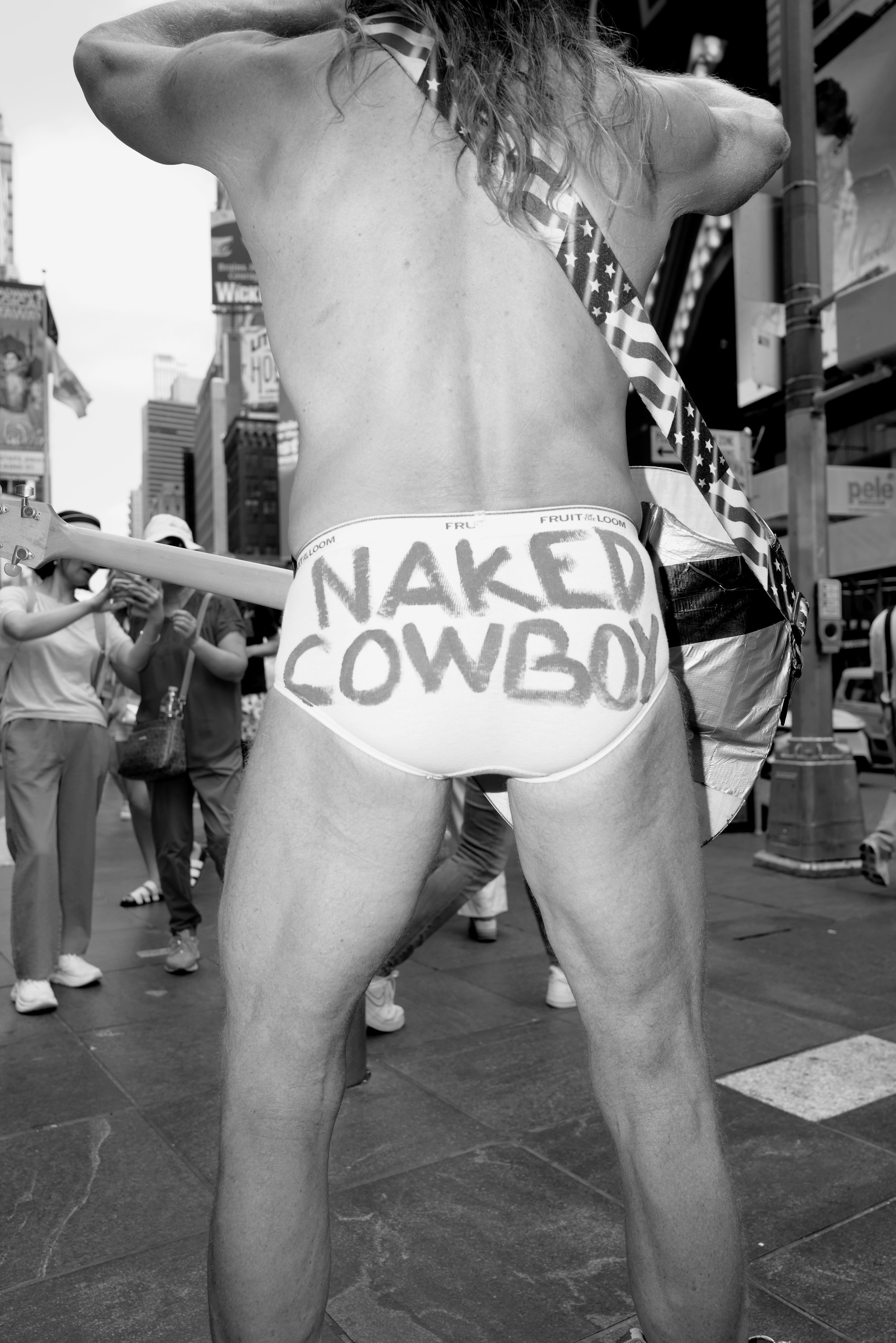 A man with no clothes, wearing only underwear with the words 'NAKED COWBOY' written on the back, standing in Times Square during a parade or event.