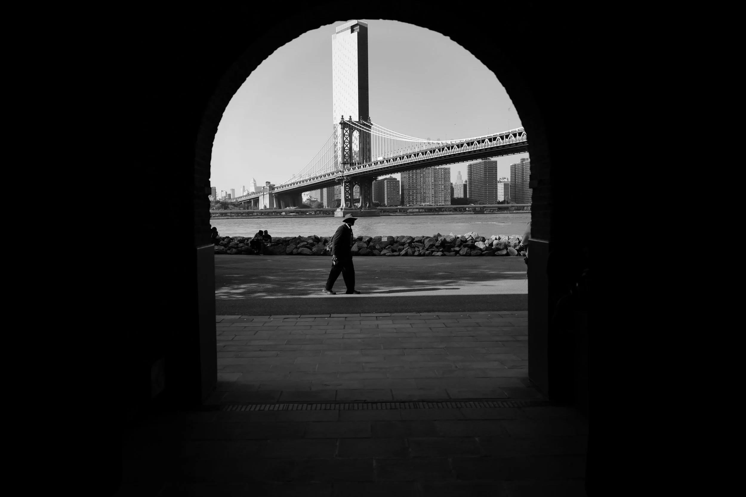 View of the Brooklyn Bridge and cityscape in New York City as seen through an archway, with a man walking on a path near the river.