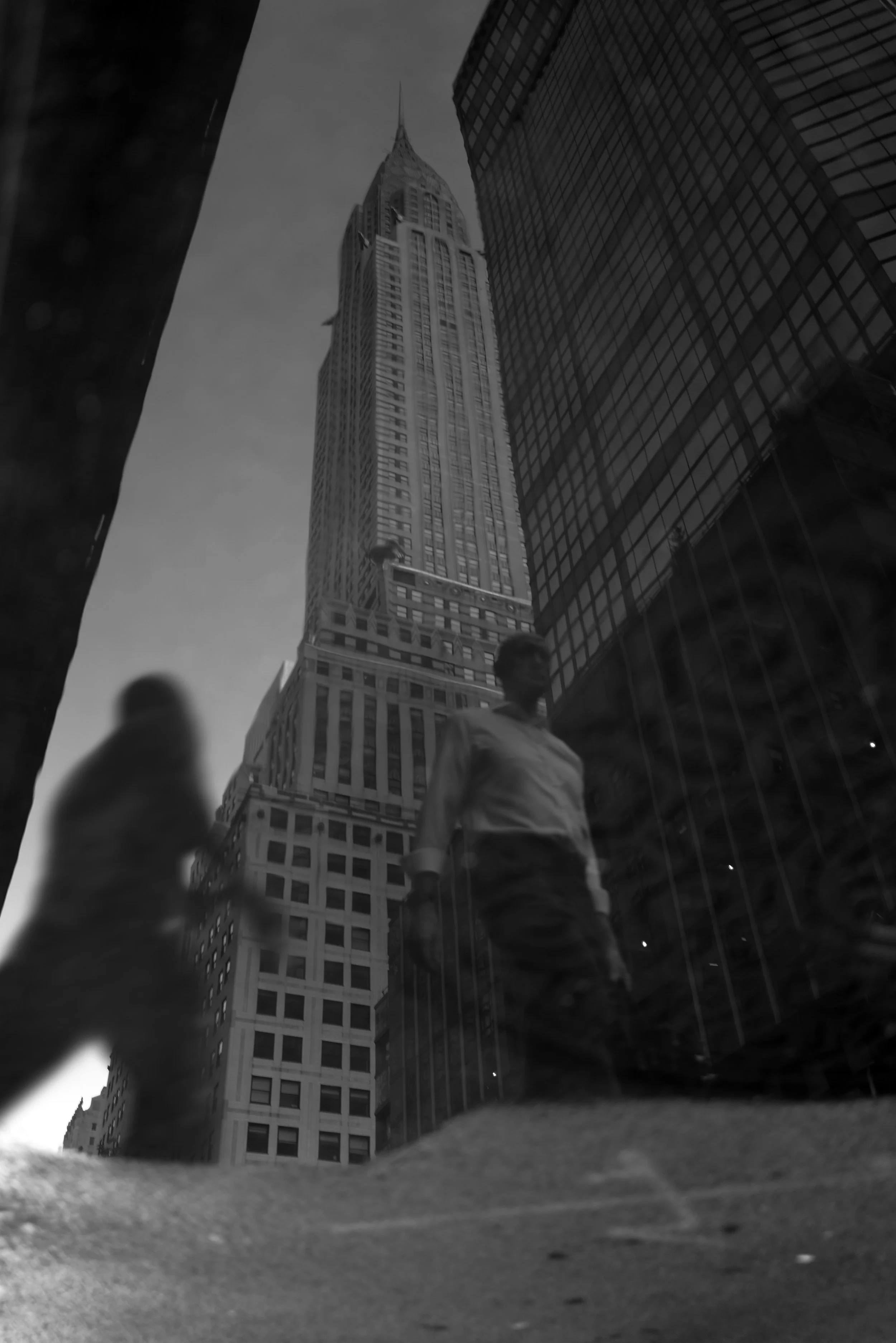 A black and white photograph of the Empire State Building in New York City, viewed from the ground with tall surrounding buildings, and reflections of pedestrians in a nearby glass surface.