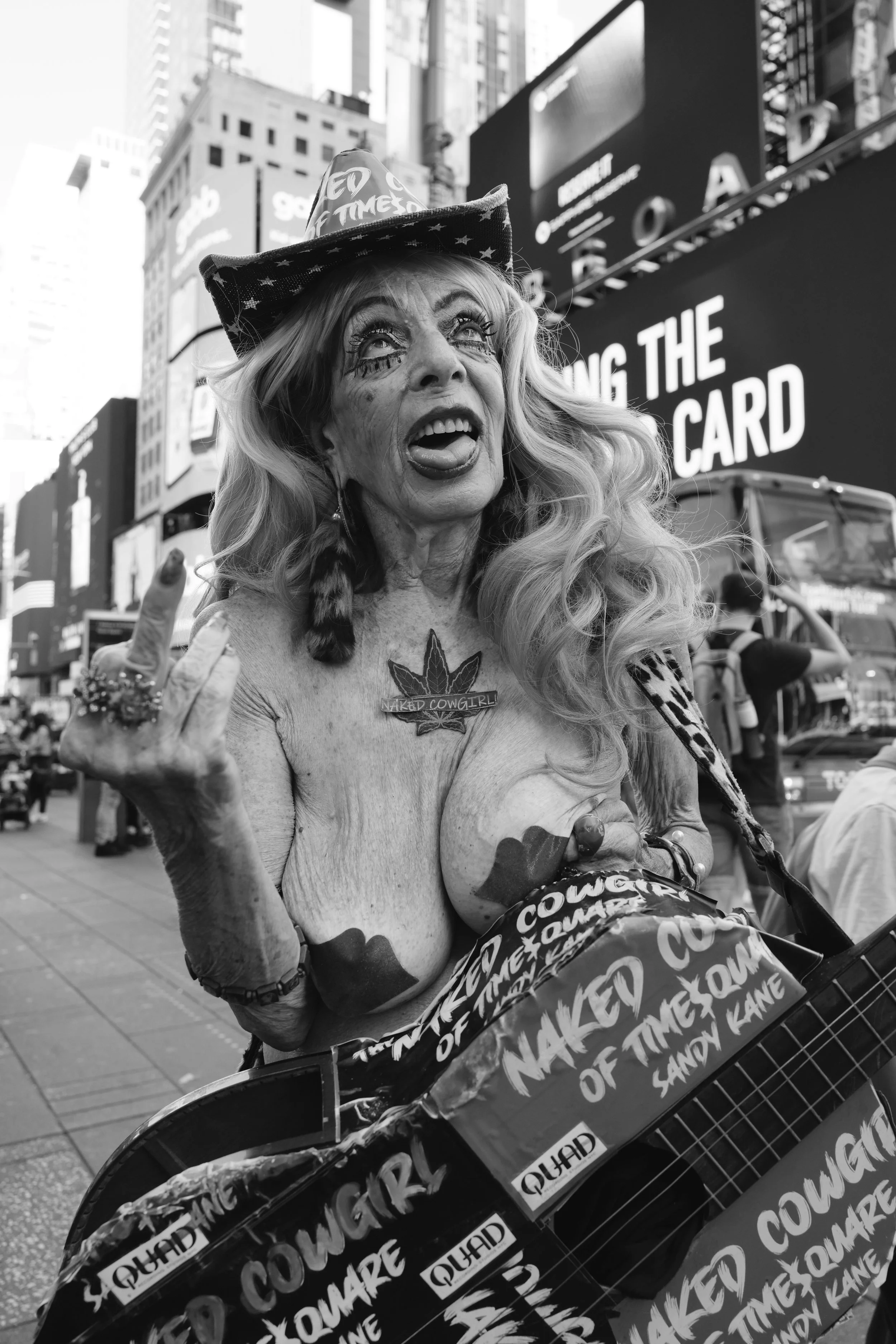 A woman with heavily makeuped face, long wavy hair, wearing a cowboy hat, and a necklace that says 'Naked Cowgirl', standing on a city street with bright billboards and a bus in the background. She is topless, with painted breasts, and is making a provocative gesture with her hand. She is mindfully expressive, with her tongue sticking out slightly.