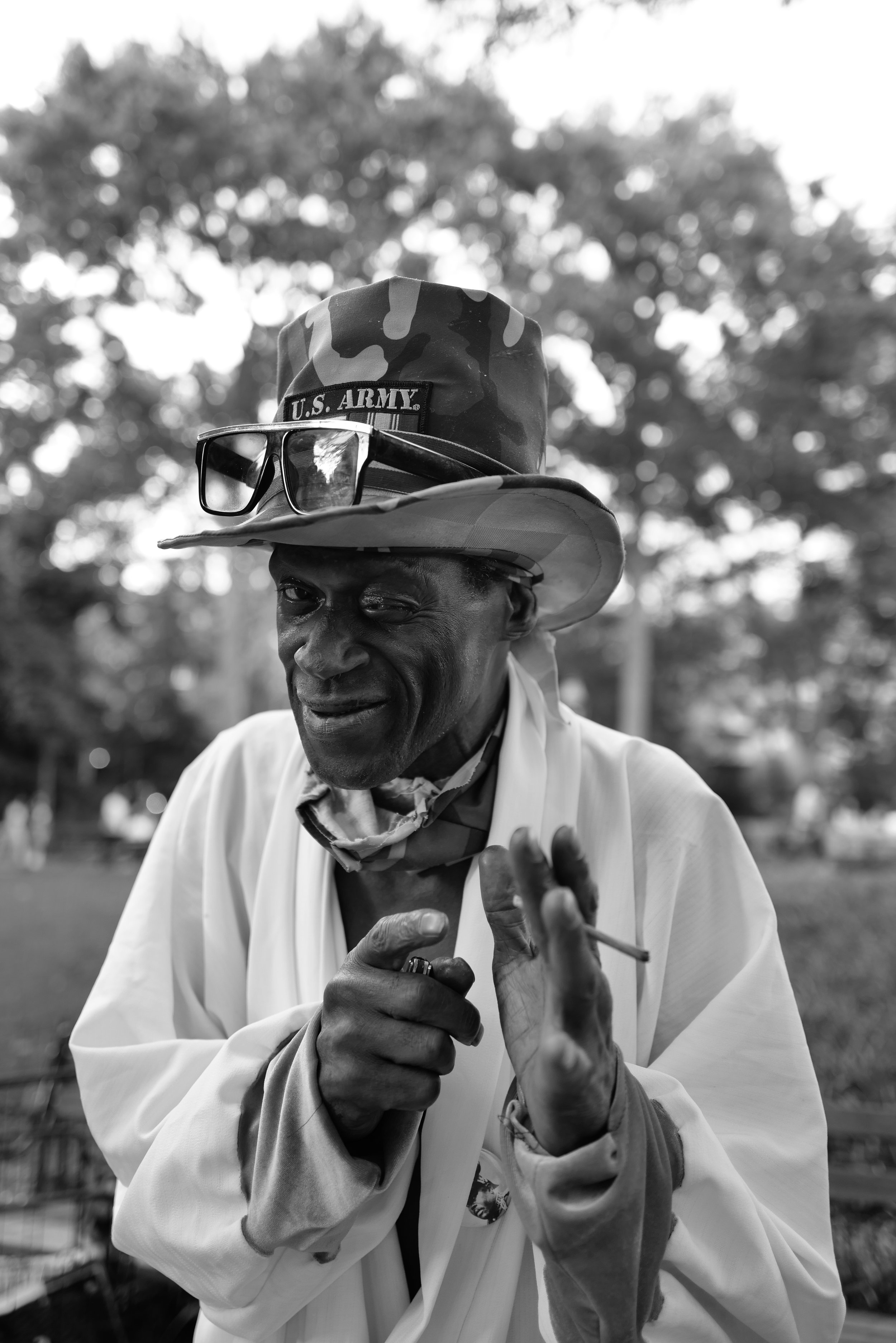 A man with dark skin wearing a white coat, a bandana, and a hat with sunglasses on top. The hat has a U.S. Army label and. The man is pointing at the camera and holds a cigarette in his left hand. The background shows outdoor trees and blurred people.