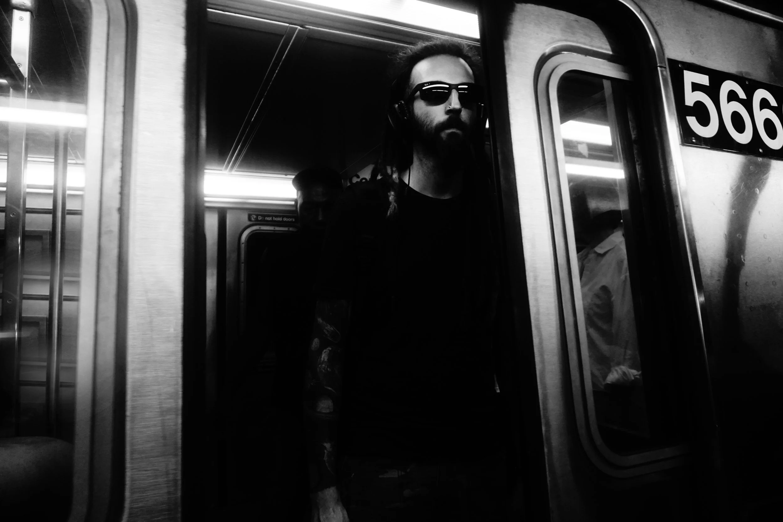 A man with a beard and sunglasses, wearing headphones, is entering a subway train through the door, with a train car number 566 visible. The photo is in black and white.