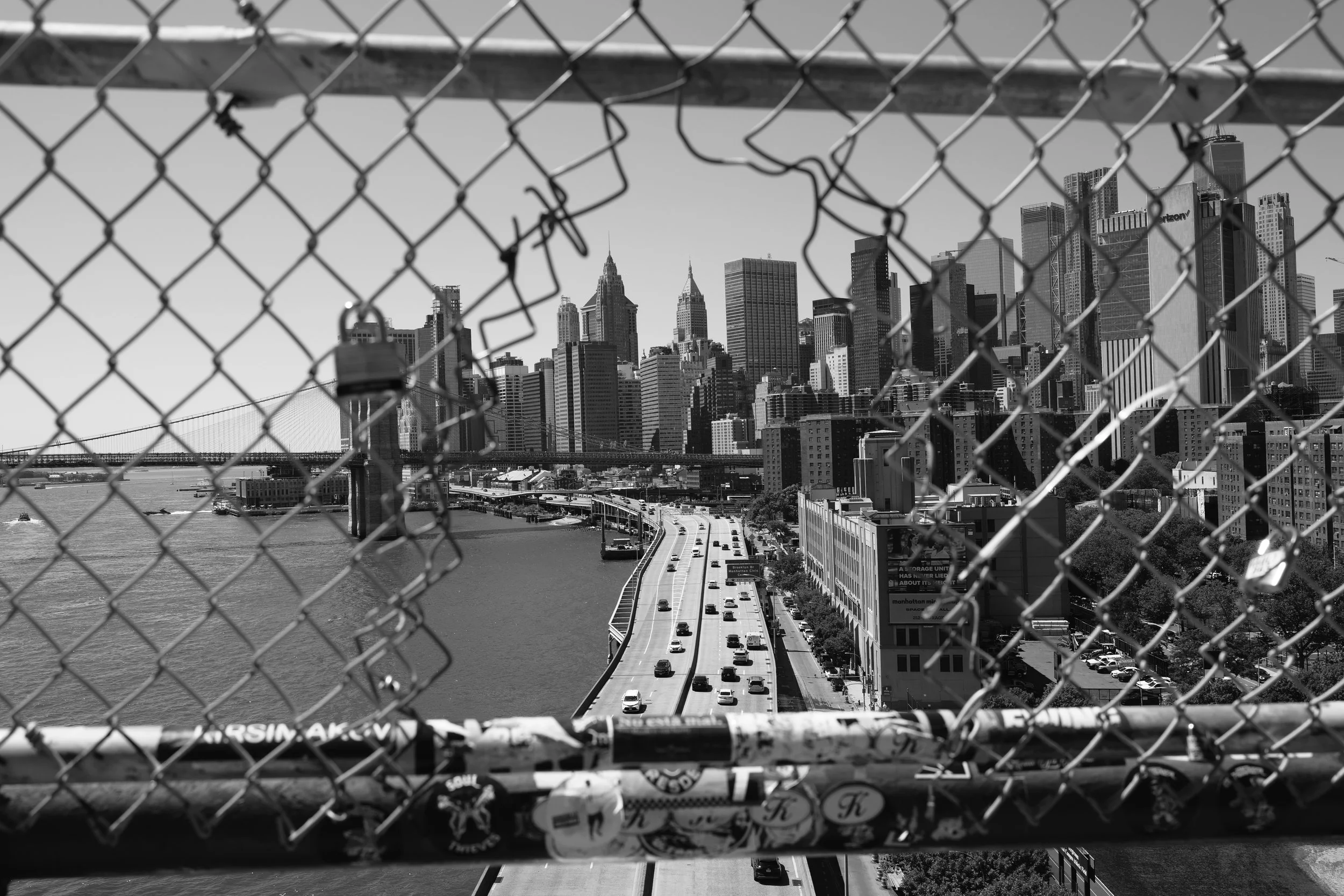 View of Manhattan skyline through a chain-link fence, with a highway and river in the foreground in black and white.