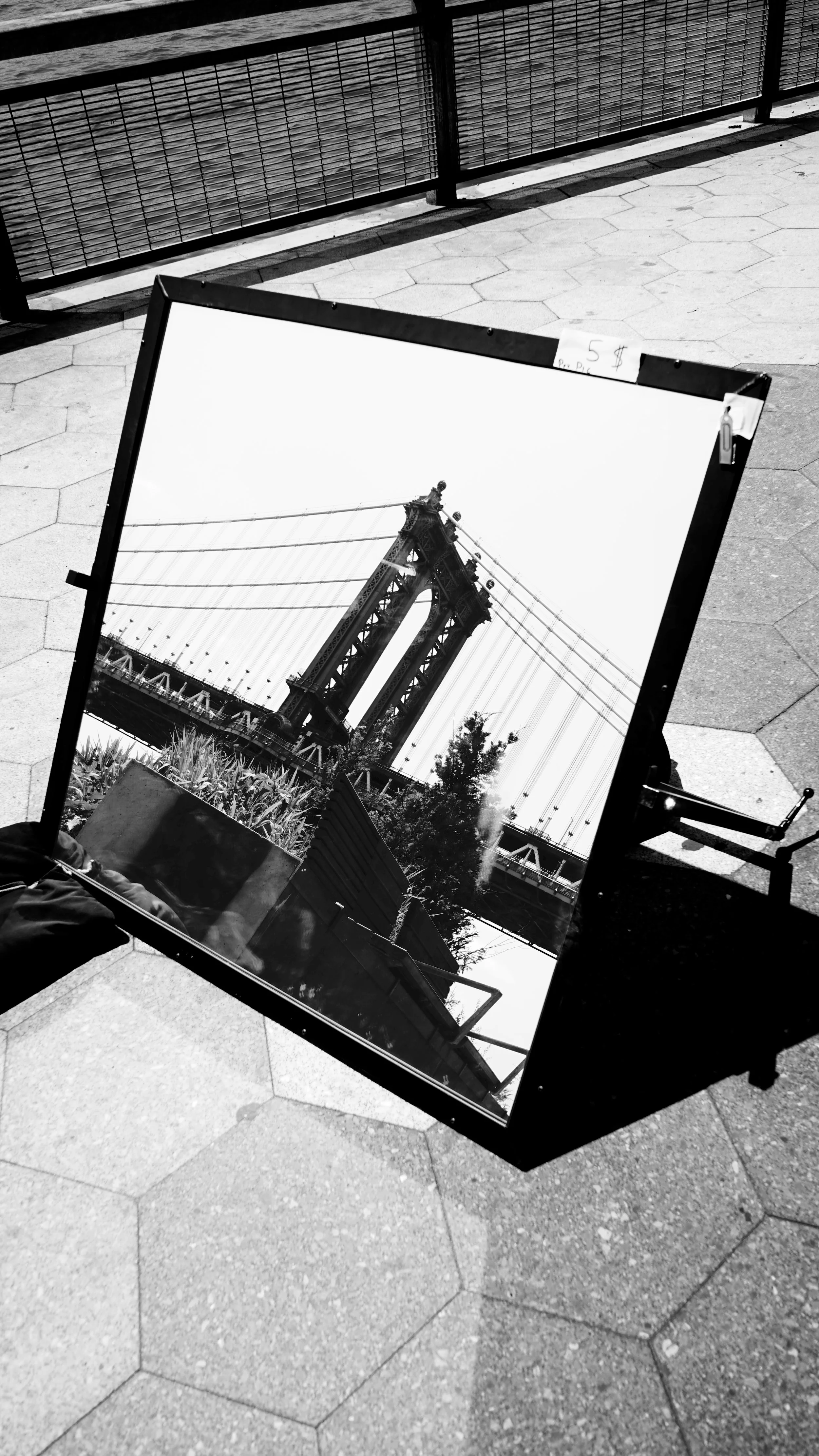 Black and white photograph of a framed picture of the Brooklyn Bridge leaning on the pavement, with a fence and hexagonal tiles in the background.